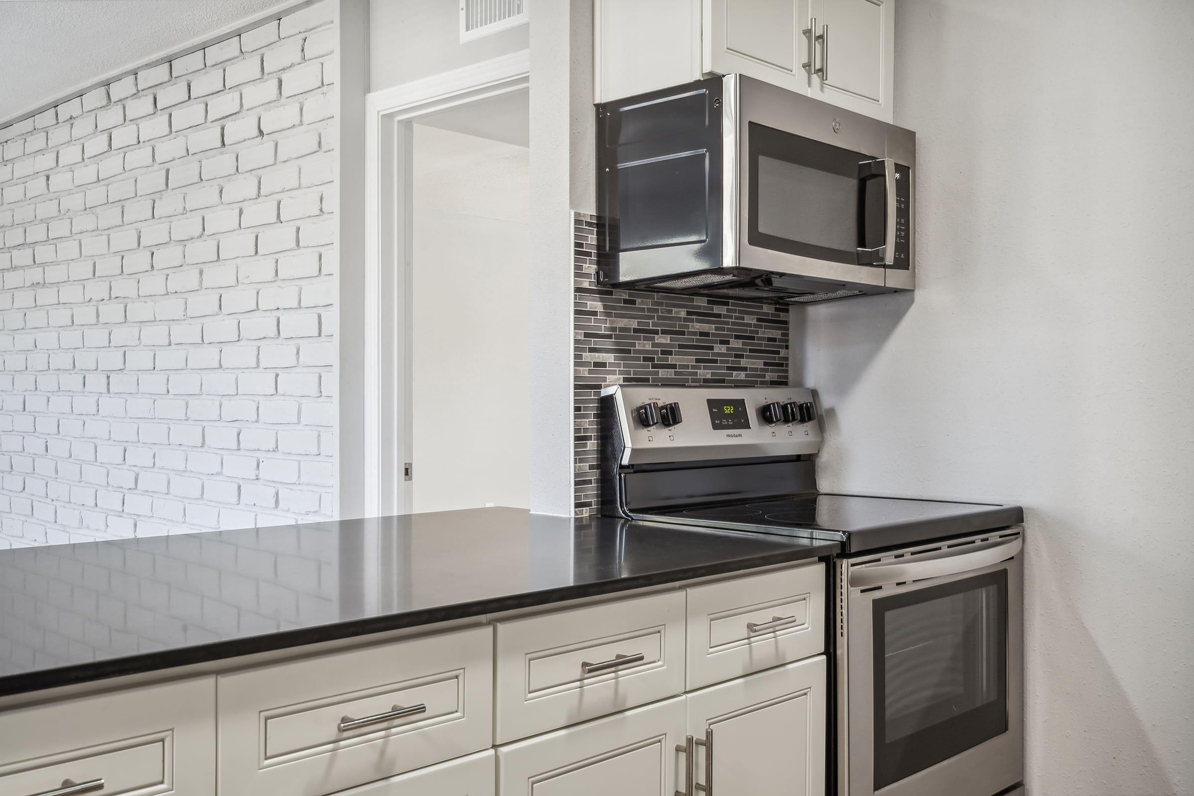 Modern kitchen featuring sleek white cabinetry, a stainless steel microwave above a black stove, and a dual oven. The countertop is dark, complementing the bright backsplash made of small tiles. A textured white brick wall adds a contemporary touch to the space.