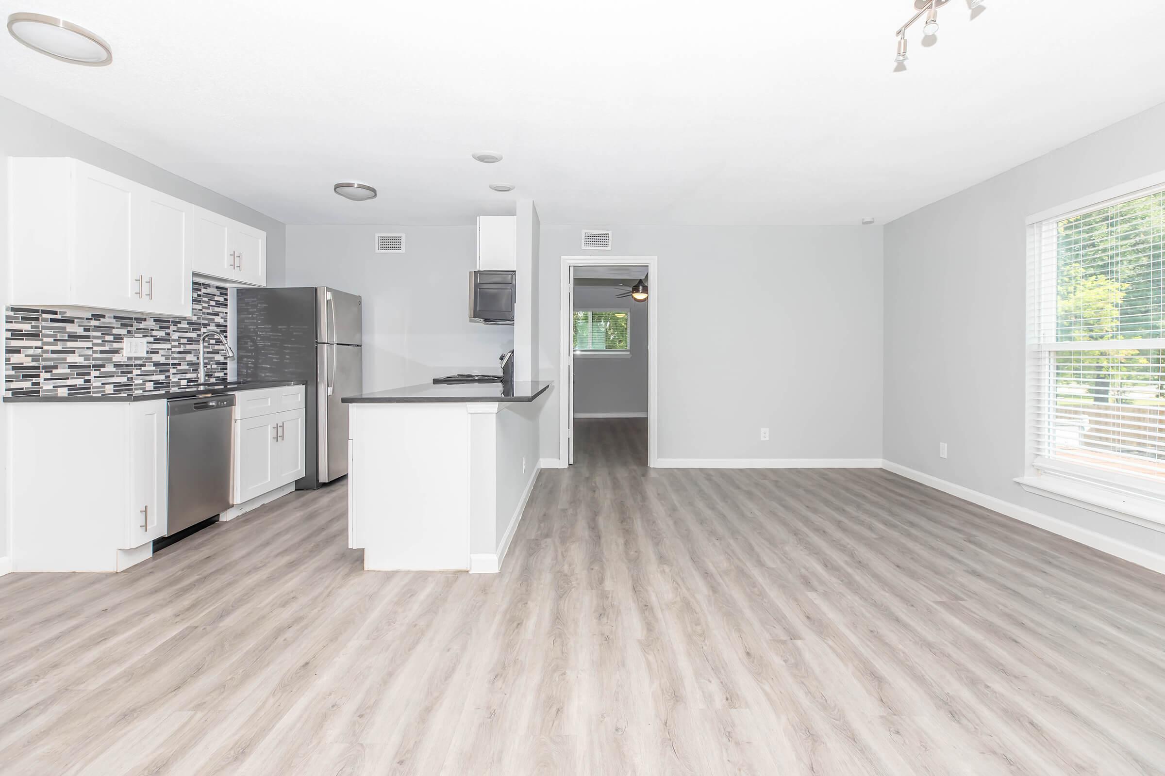 Spacious open-concept living area featuring light gray walls and wood-style flooring. The kitchen area includes modern white cabinets, stainless steel appliances, and a black countertop. Large windows allow natural light to fill the room, creating a bright and inviting atmosphere.