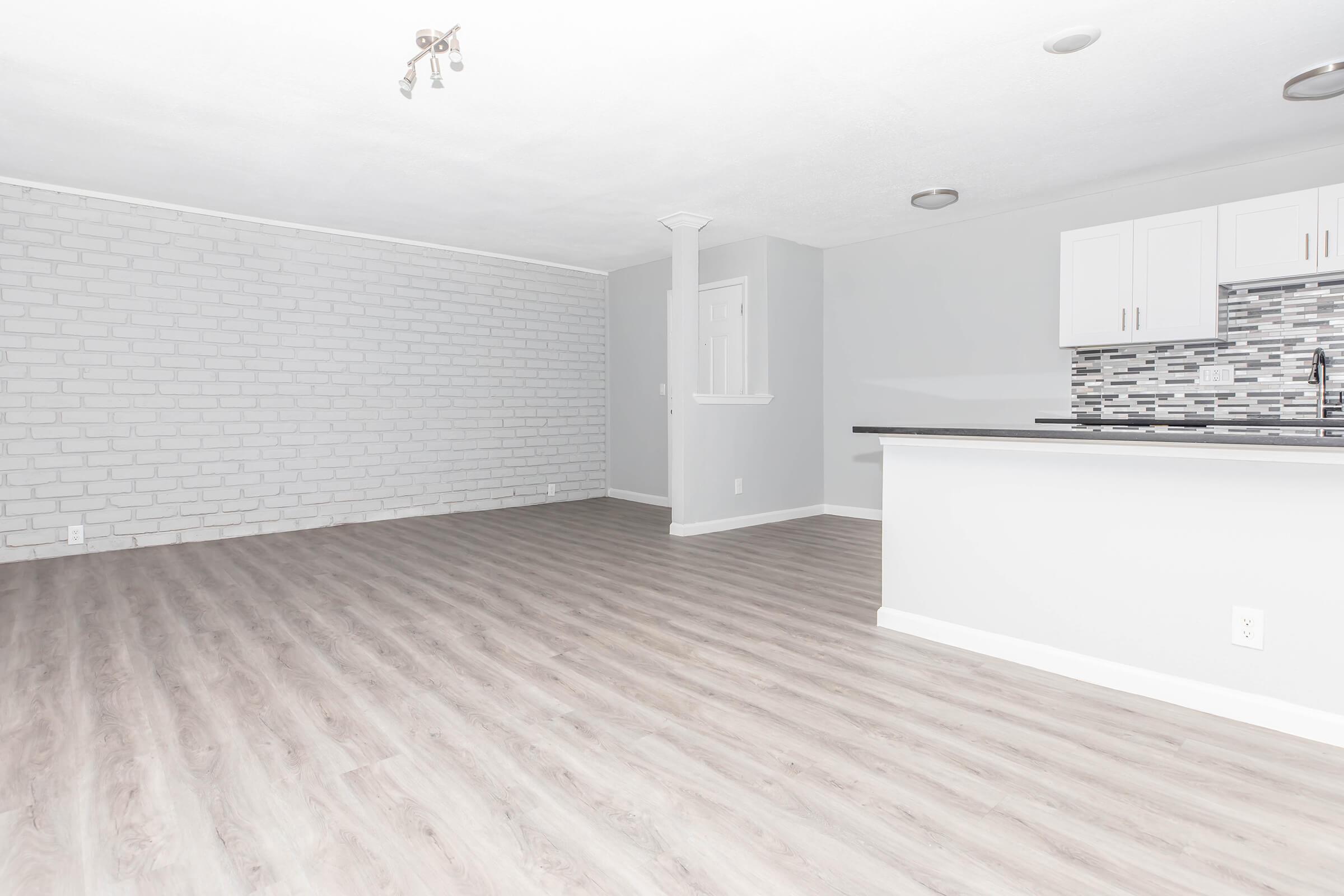 A spacious, empty room featuring light-colored walls, a brick accent wall, and modern laminate flooring. The kitchen area is visible with white cabinets and a tiled backsplash, while natural light floods the space. The room has a clean, minimalist aesthetic, ideal for various furnishings.