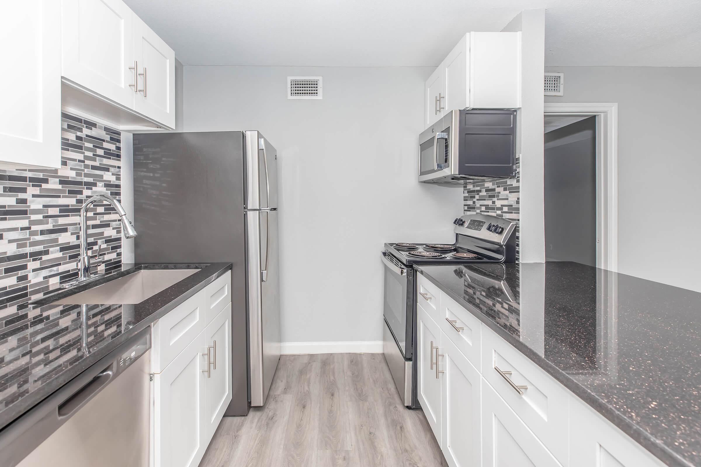 Modern kitchen featuring stainless steel appliances, including a refrigerator, oven, and microwave. The cabinetry is white with sleek handles, and the backsplash consists of dark and light tiles. A dark countertop contrasts with the light walls and flooring, creating a stylish and contemporary look.
