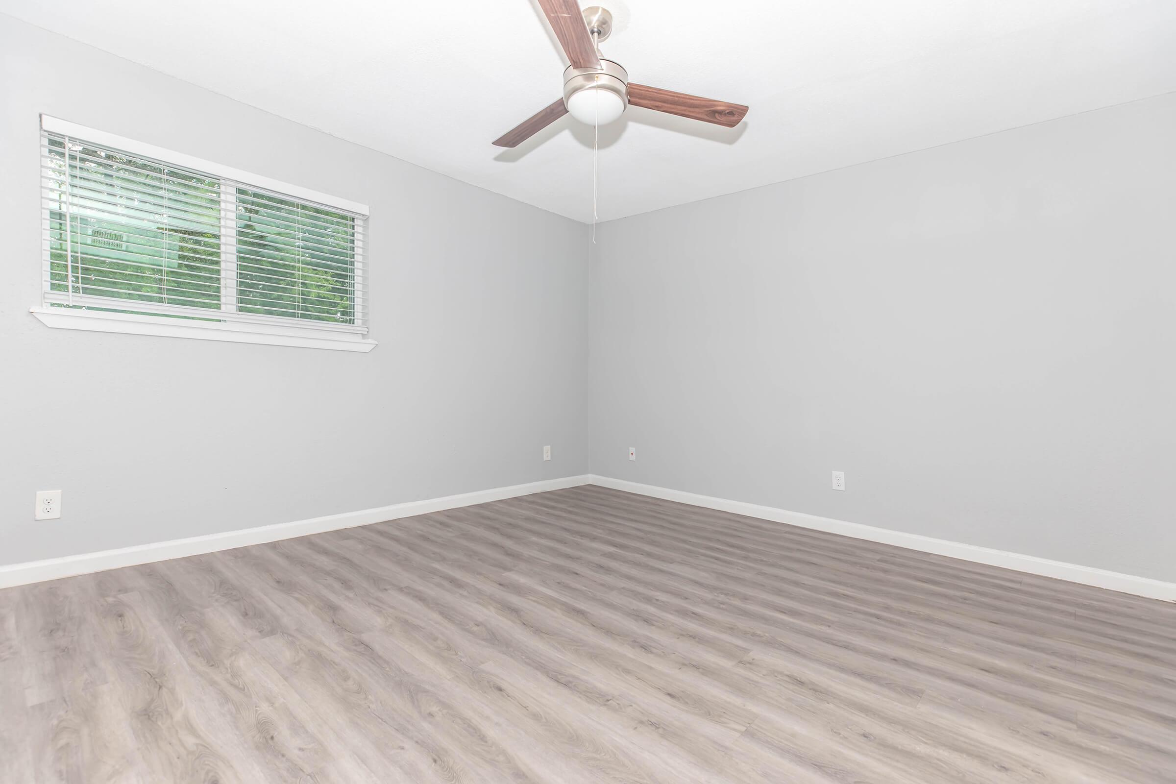 Empty room with light gray walls and a ceiling fan. A window with white blinds allows natural light in. The floor is covered with light-colored laminate flooring, creating a spacious and minimalist atmosphere.