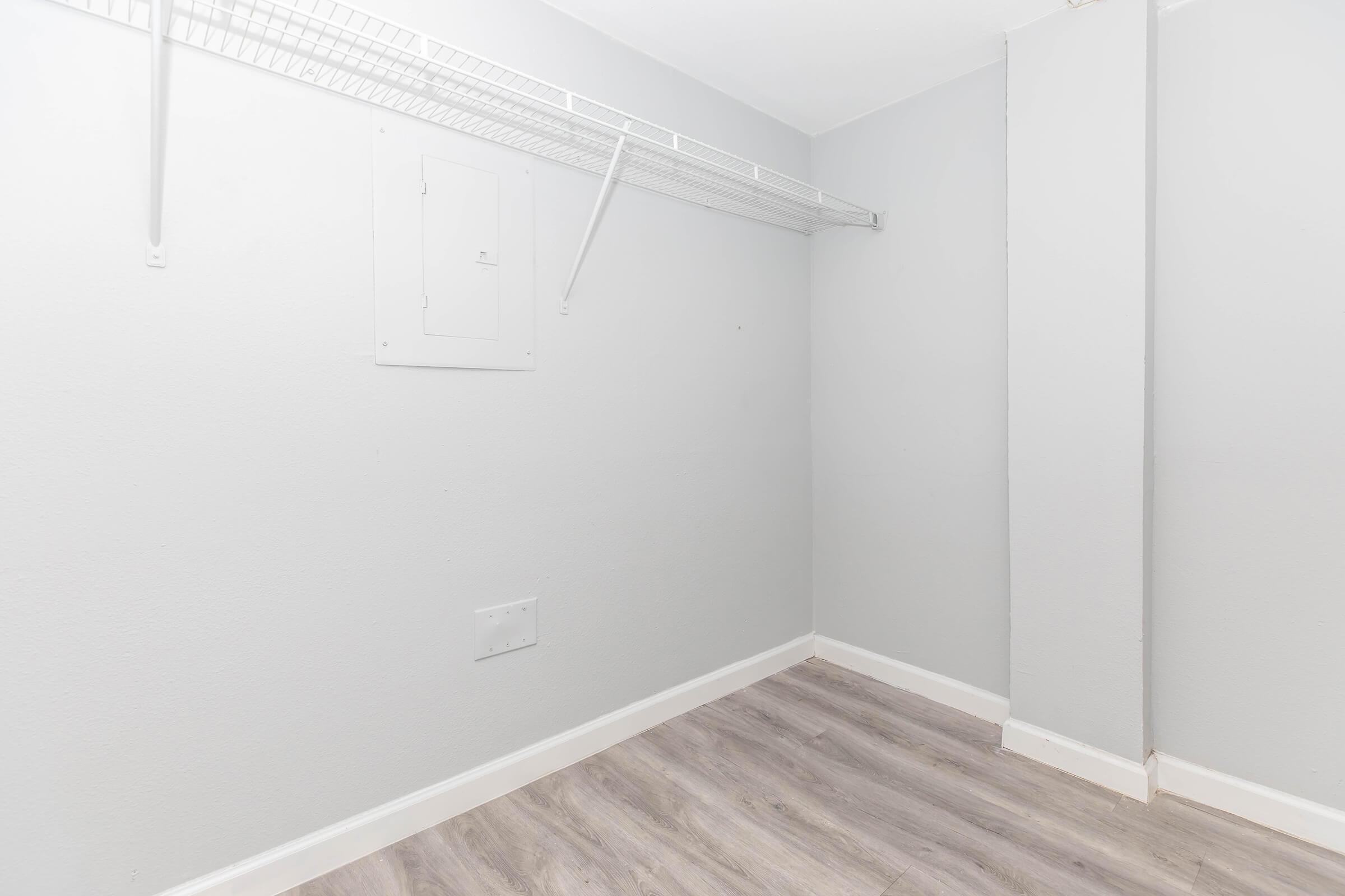Empty closet with light gray walls and a light wood laminate floor. A wire shelf runs along the top, and there is a small access panel on the wall. The space is well-lit and minimalistic, suitable for storage.