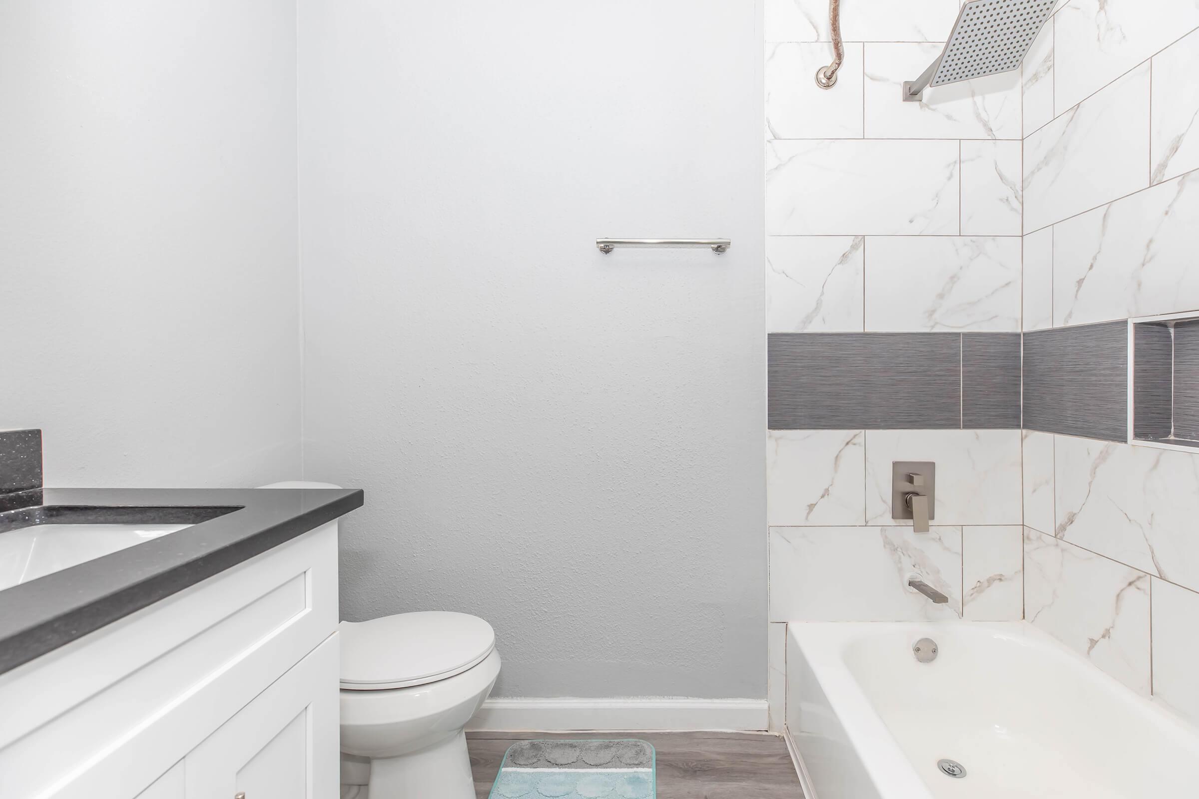 A modern bathroom featuring a white vanity with a dark countertop, a toilet, and a bathtub with a shower. The walls are tiled with light marble and gray accents. A towel rack is mounted on the wall, and a soft bathmat lies on the floor. The overall color scheme is neutral and bright.