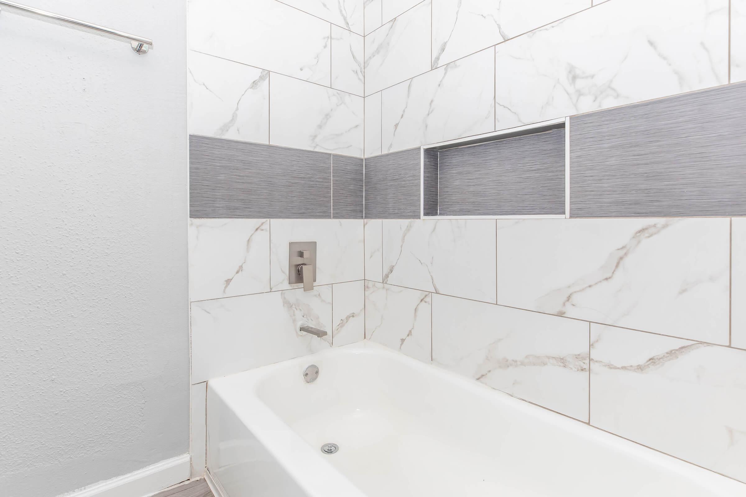 A modern bathroom with a white bathtub and elegant wall tiles. The tiles feature a combination of white marble with grey accents, and there is a small recessed shelf in the wall. A minimalist faucet is visible next to the tub, adding to the contemporary design.
