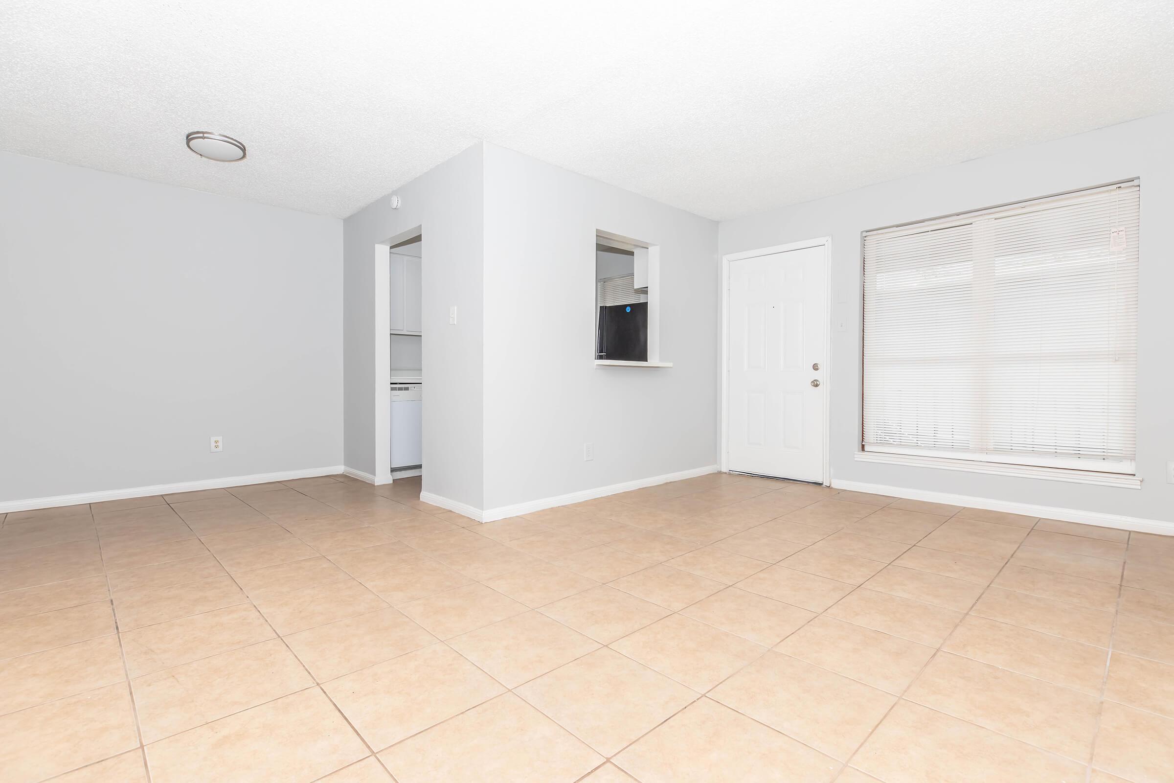 Spacious, empty living room with light-colored walls and tiled flooring. Features a large window with blinds, a front door, and an open entrance to a small kitchen area. The overall ambiance is bright and inviting, with natural light illuminating the space.
