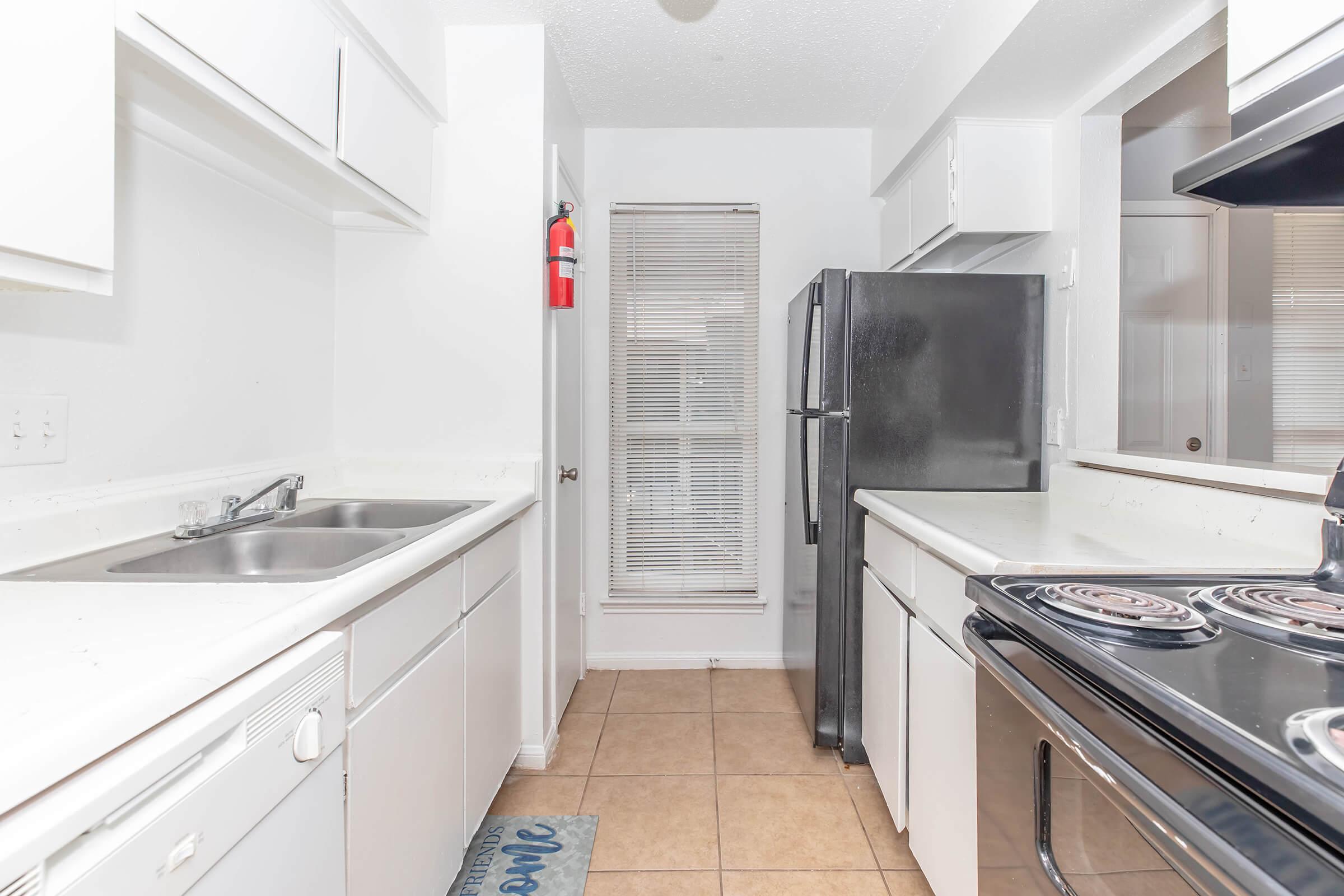 A bright, clean kitchen featuring white cabinets, a black refrigerator, and a black stove. There are dual sinks with a window above, a fire extinguisher mounted on the wall, and tiled flooring. Natural light enters from a window near the back, enhancing the spacious feel of the area.