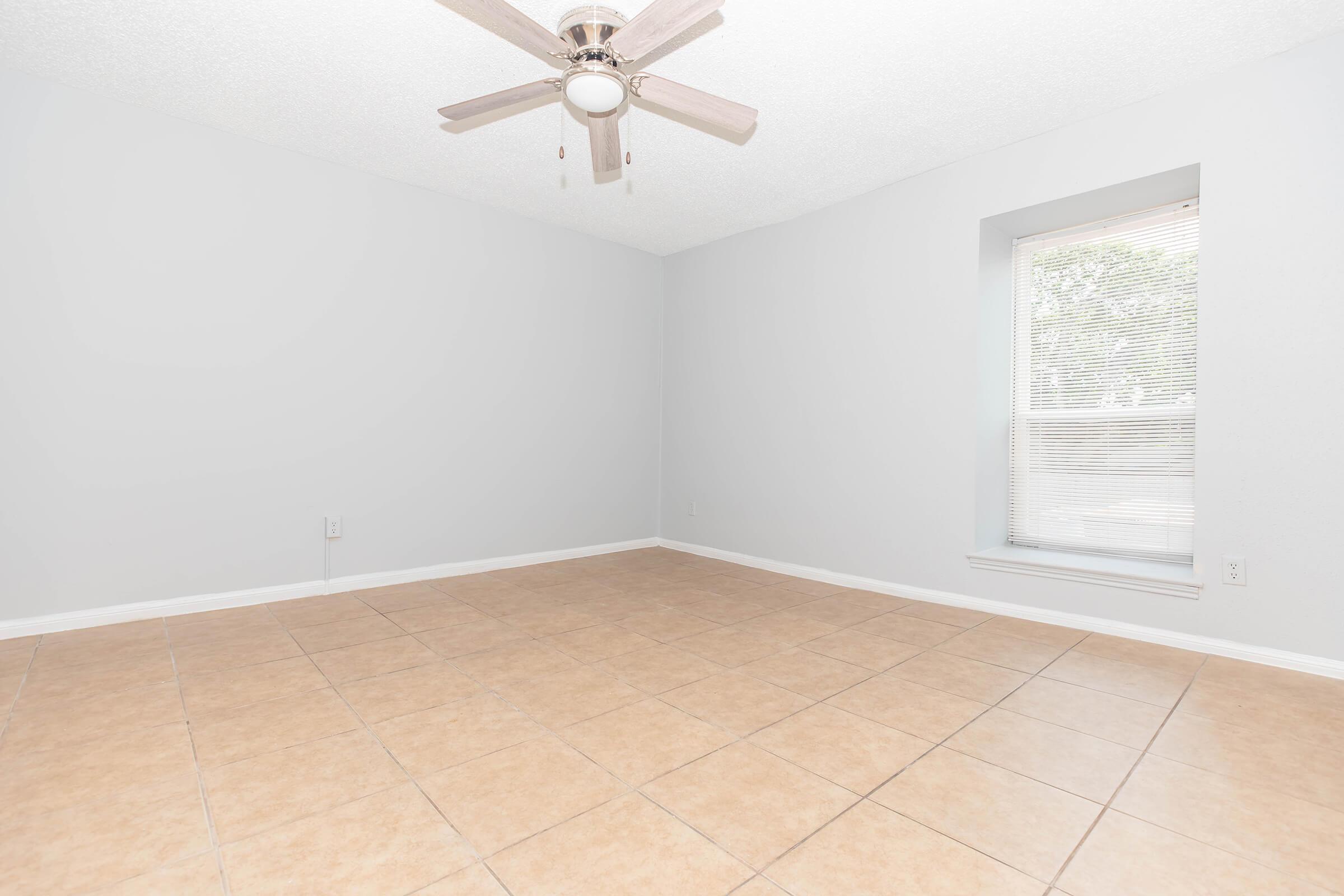Empty room with light gray walls, a ceiling fan, and beige tile flooring. A window with blinds allows natural light to enter. The space appears clean and uncluttered, suitable for various uses or decoration.