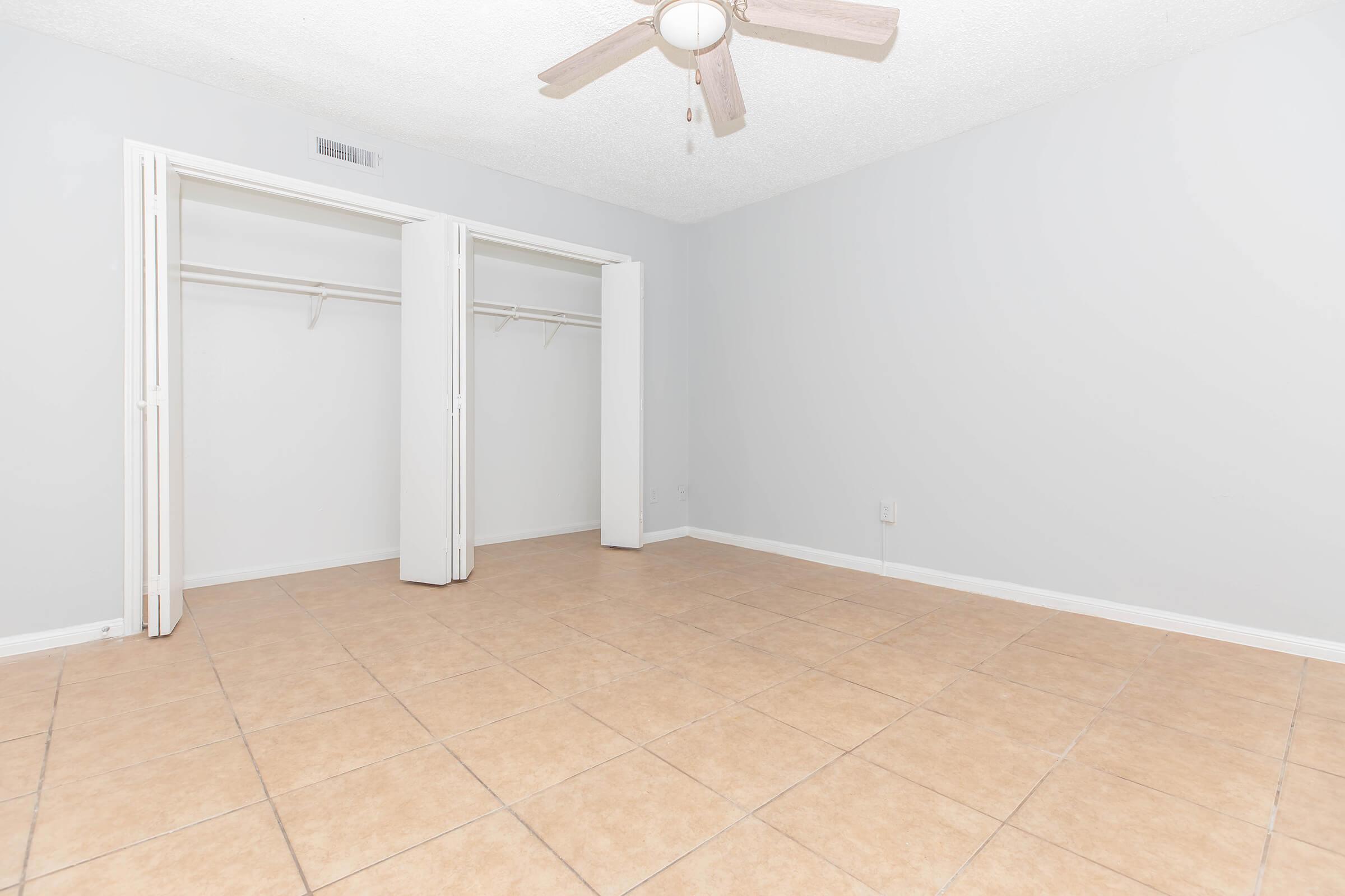 A clean, empty room with light gray walls and a ceiling fan. The floor is tiled with beige tiles. There are two white sliding closet doors on one wall, both open, revealing empty closet spaces. The room appears spacious and well-lit, with a simple and minimalist design.