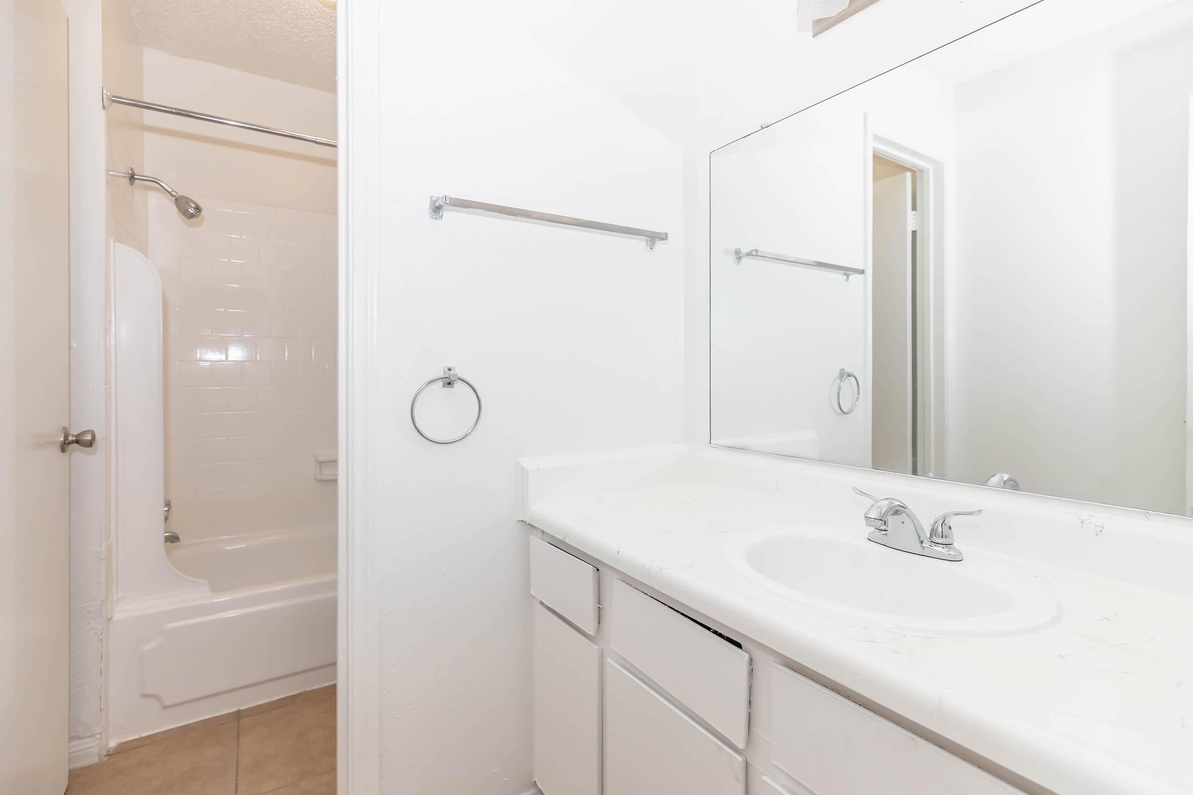 A clean and simple bathroom featuring a bathtub with a showerhead above, a large mirror, and a double sink vanity. The walls are painted white, and the floor has light tiles, creating a bright and open atmosphere.