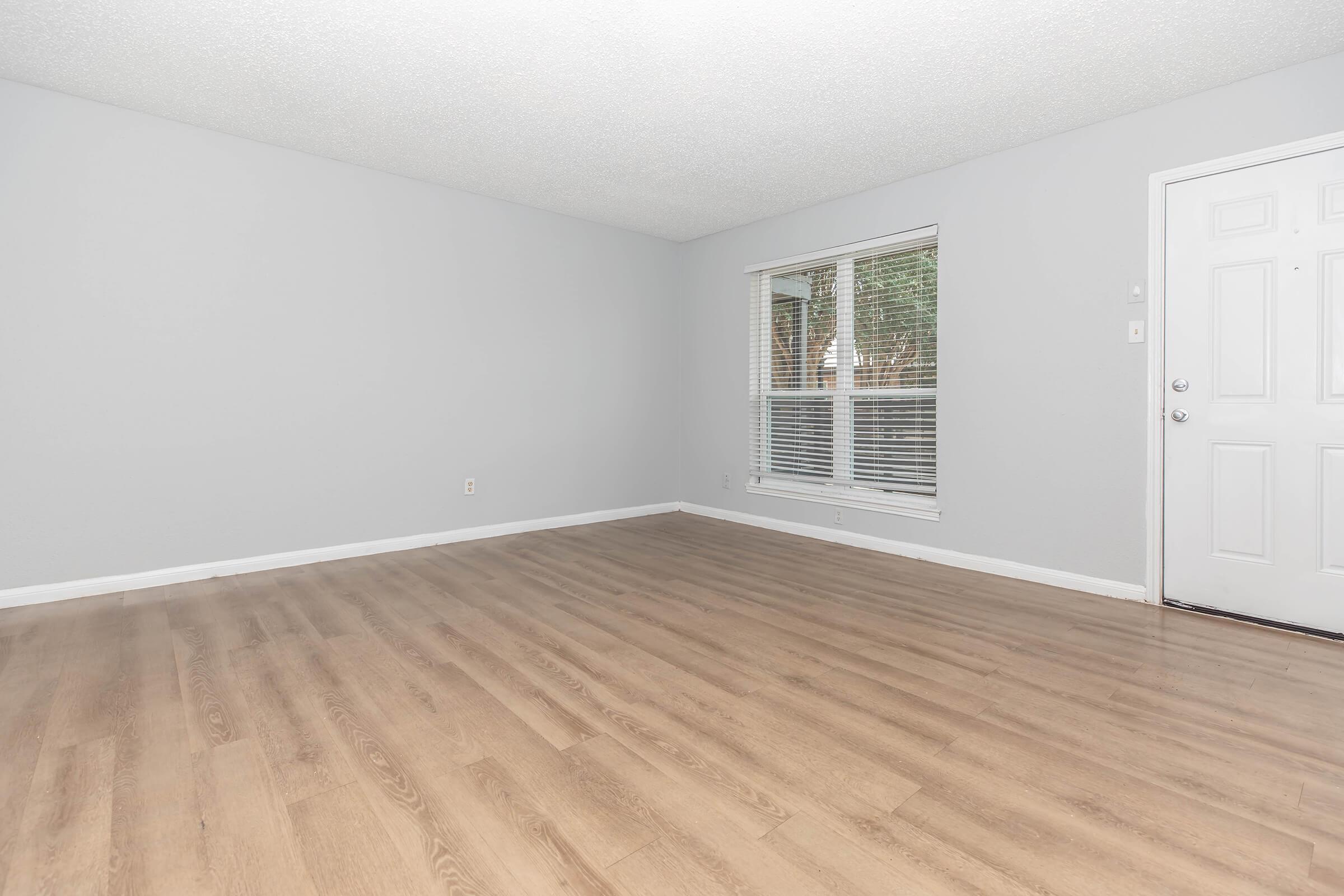 A spacious, empty living room featuring light gray walls and laminate flooring. Large windows allow natural light to stream in, and a white front door is visible on the right. The room is devoid of furniture, creating an open and airy atmosphere.