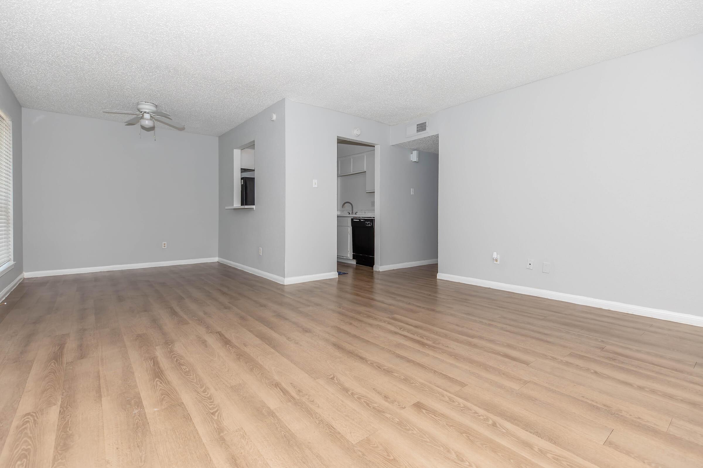 A spacious, light gray living room featuring a ceiling fan, hardwood-style flooring, and an open layout. The room is empty, with a small opening leading to a kitchen area visible in the background. Natural light comes through a window, enhancing the airy feel of the space.