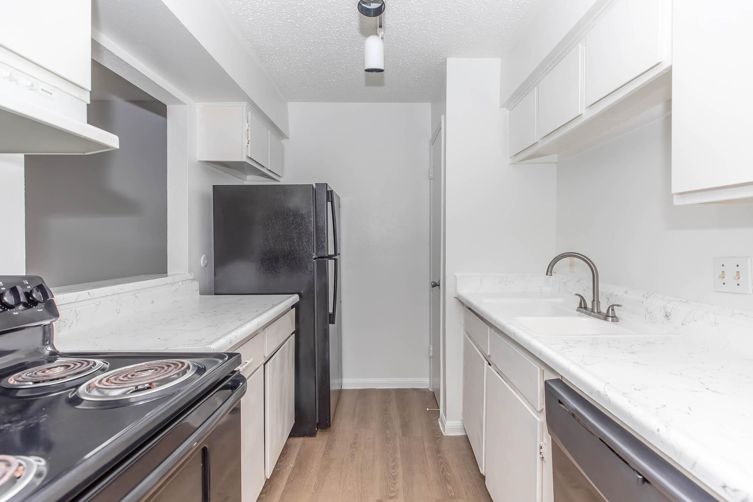 Modern kitchen featuring white cabinets, granite-style countertops, a black stove, and a refrigerator. The space is well-lit, showcasing a minimalist design with a neutral color palette and wooden flooring. A sink is positioned near the window, enhancing the functionality of the layout.