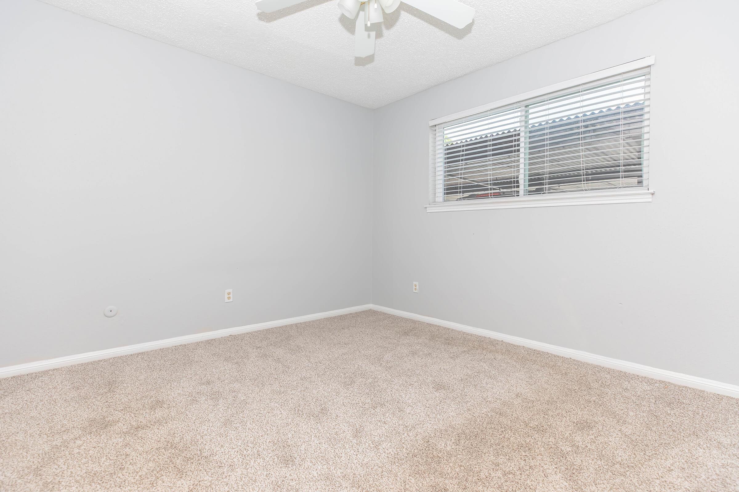 A plain, empty bedroom with light gray walls and carpeted flooring. There is a ceiling fan and a window with blinds letting in natural light. The room is devoid of furniture and decorations, creating a spacious and minimalistic atmosphere.