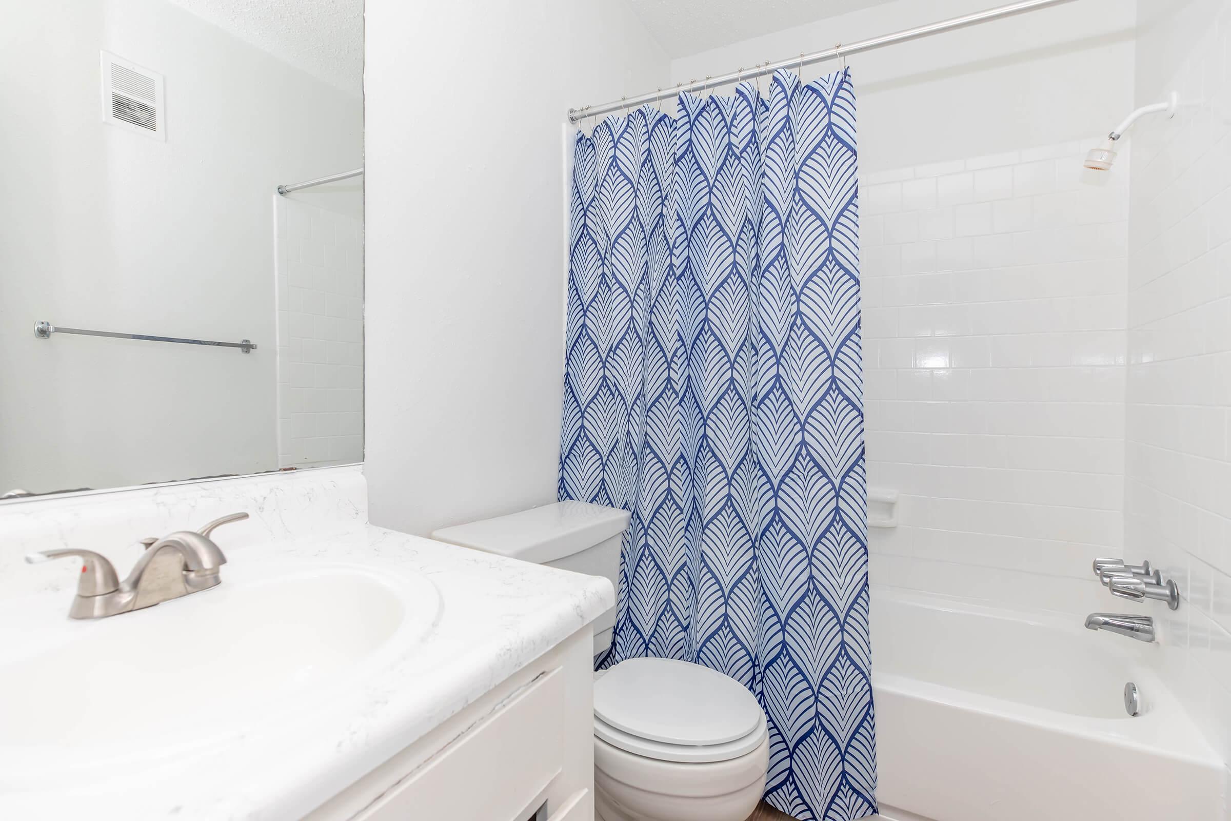 A modern bathroom featuring a white countertop, a toilet, and a bathtub with a white tile surround. A blue and white patterned shower curtain adds color to the space. There's a mirror on the wall, and the walls are painted white, creating a clean and bright atmosphere.