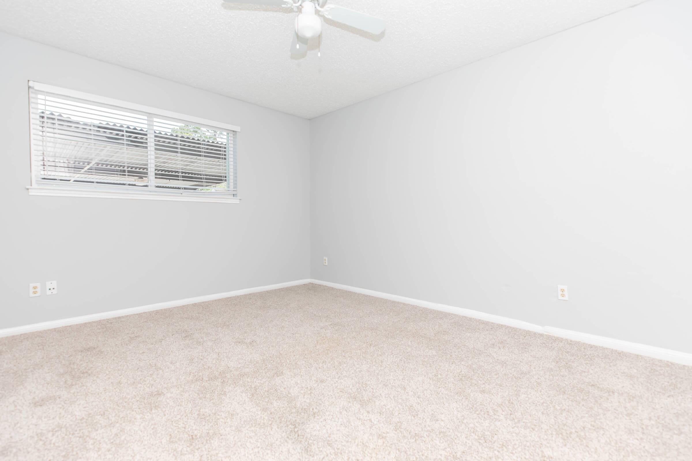 A spacious, empty room with light gray walls and beige carpet. A window with blinds on one wall allows natural light to enter. A ceiling fan is installed, adding a modern touch. The room has minimal furnishings, creating a clean and open atmosphere, suitable for various uses.