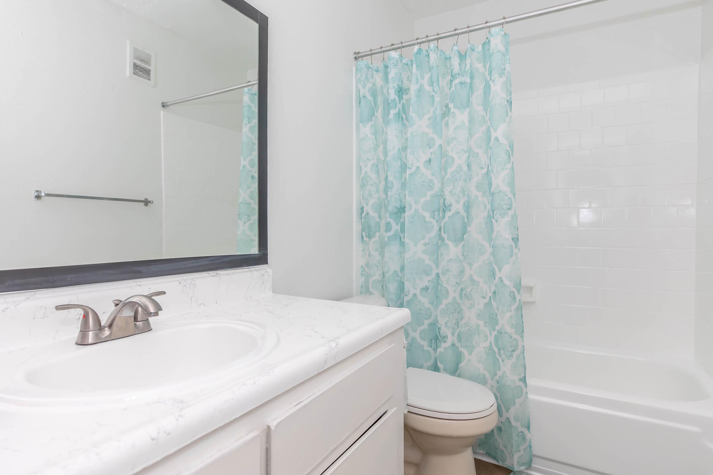 A clean and modern bathroom featuring a white sink with a marble countertop, a mirror above the sink, a light blue patterned shower curtain, and a bathtub with a white tile surround. The space is well-lit and has neutral-colored walls. A towel rack is mounted on the wall.