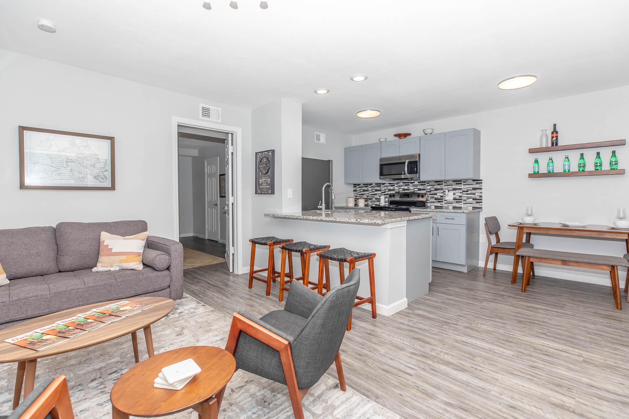 A modern living space featuring a cozy gray sofa, a round wooden coffee table with board games, and a kitchen area with gray cabinets and a marble countertop. Stools at the kitchen island complement the decor, while a small dining table is positioned against a wall with decorative bottles. Bright, neutral colors create a welcoming atmosphere.