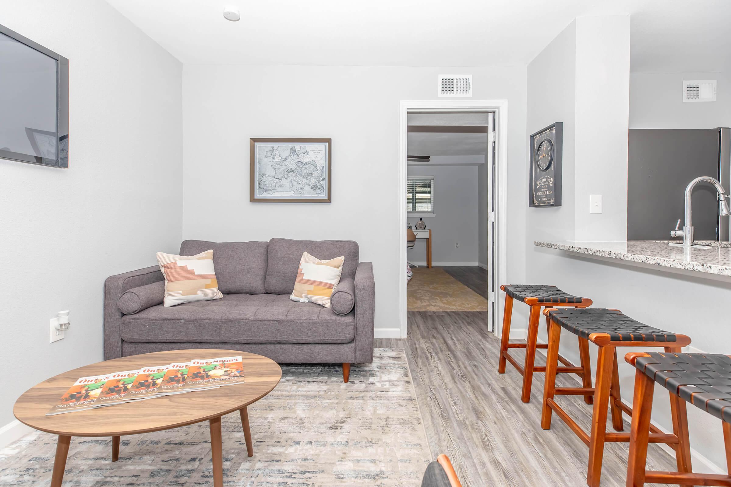 A cozy living area featuring a gray sofa with decorative pillows, a round coffee table, and a kitchenette with a breakfast bar. Three wooden stools with black seats are positioned at the bar. The walls are painted light gray, and a decorative map is framed and hung above the sofa, adding a touch of character to the space.