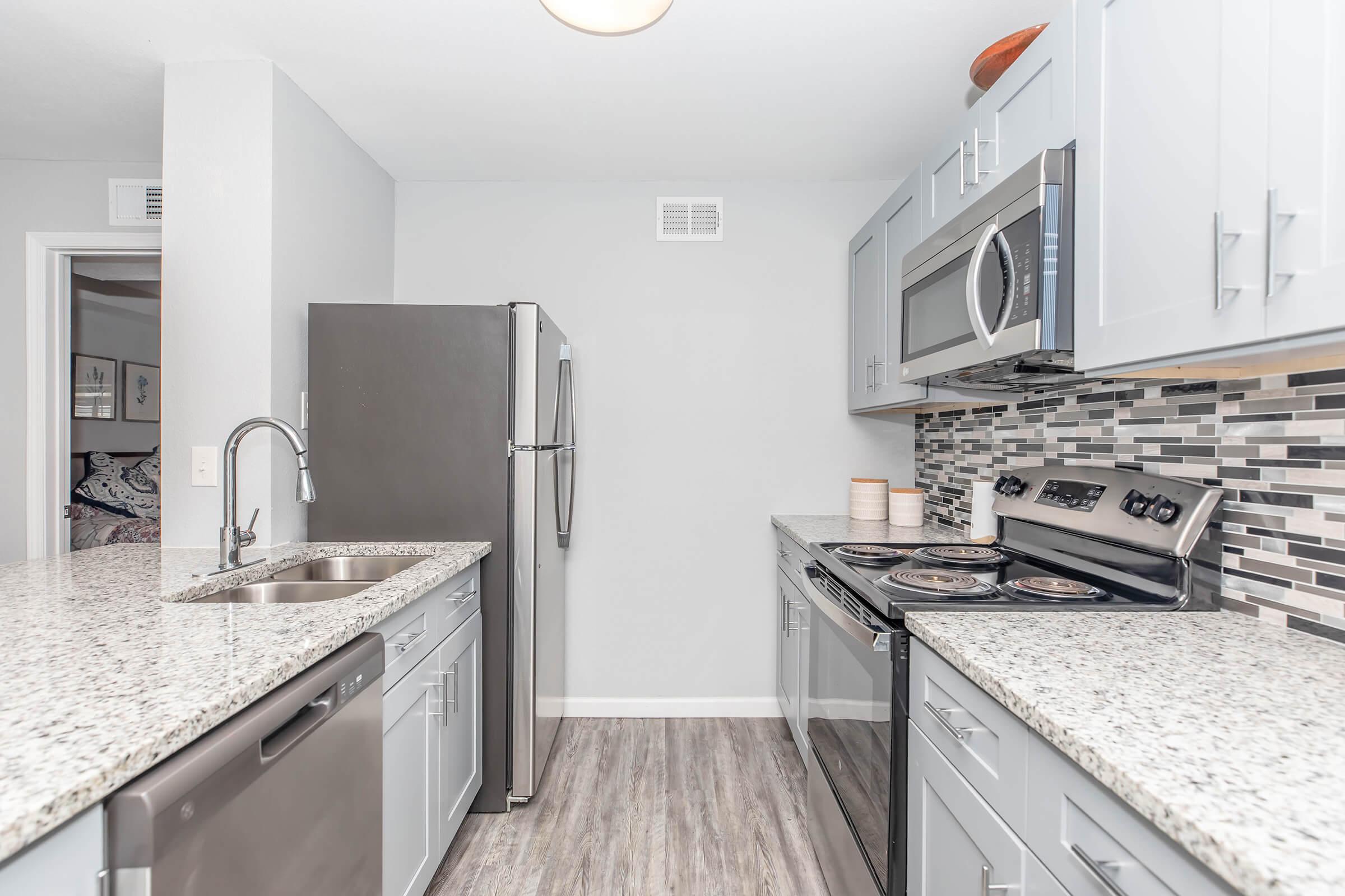 Modern kitchen with light gray cabinets, stainless steel appliances including a refrigerator, dishwasher, and stove, and a granite countertop. A mosaic backsplash adds texture. The space is well-lit and has light wood-like flooring, creating a clean and inviting atmosphere.