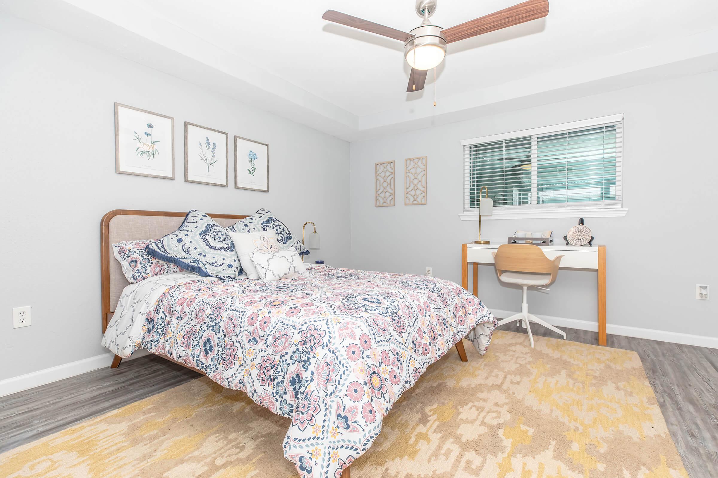 Cozy bedroom featuring a neatly made bed with a colorful patterned duvet, two decorative pillows, a wooden headboard, and a ceiling fan. A small desk with a chair is positioned nearby, alongside decorative wall art and a window with light curtains, creating a bright and inviting atmosphere.