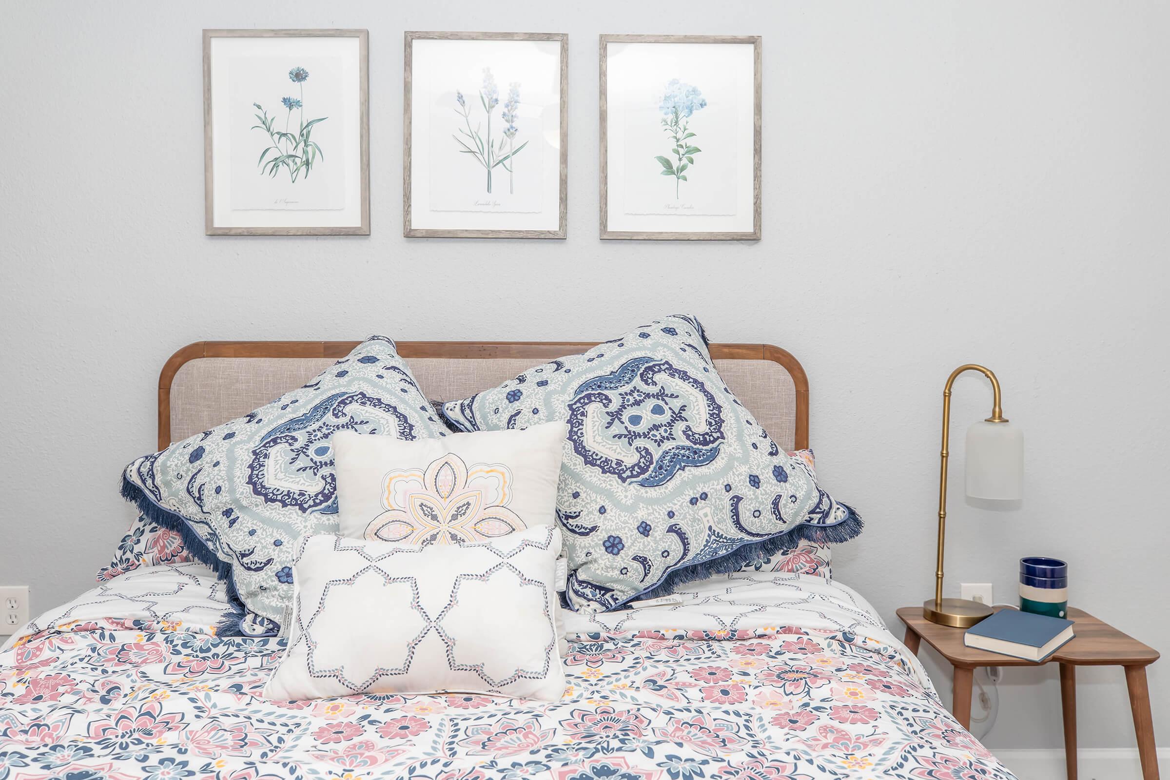A cozy bedroom featuring a wooden headboard with decorative pillows in blue and white patterns. The bed is adorned with a floral blanket. Above the headboard are three framed botanical prints. A bedside table holds a lamp and a small stack of books, adding to the inviting atmosphere.