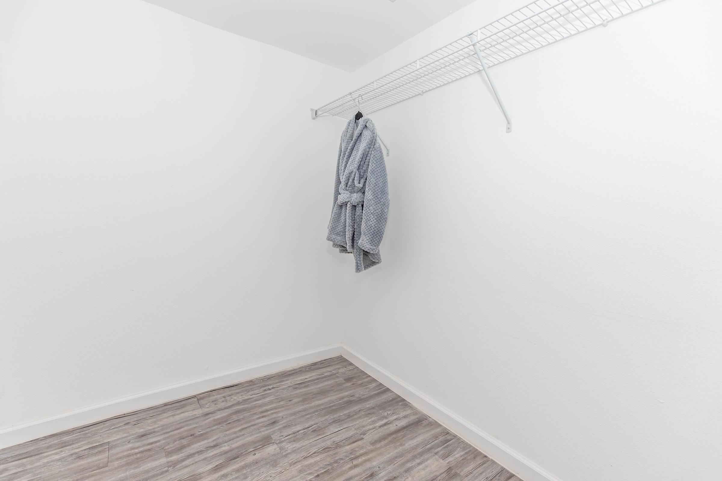 A sparsely furnished closet featuring a single wire shelf and a bathrobe hanging from a hook. The walls are plain white, and the floor is made of light-colored wood laminate, creating a clean and minimalistic look. The space is well-lit and uncluttered.