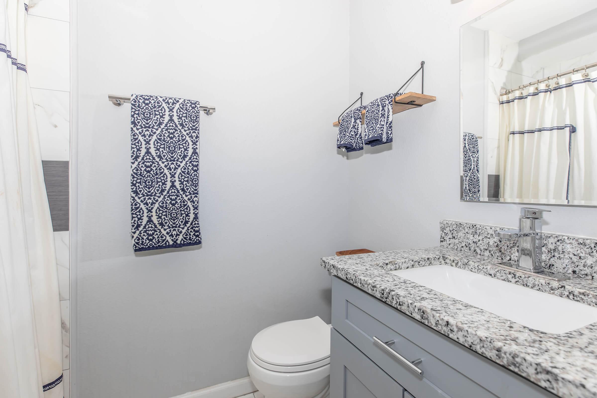 A modern bathroom with light gray walls, a white toilet, and a granite countertop. Two blue patterned towels hang on a towel rack and a shelf, while a mirror reflects the space above the sink. A beige shower curtain adds contrast to the decor, creating a clean and stylish appearance.