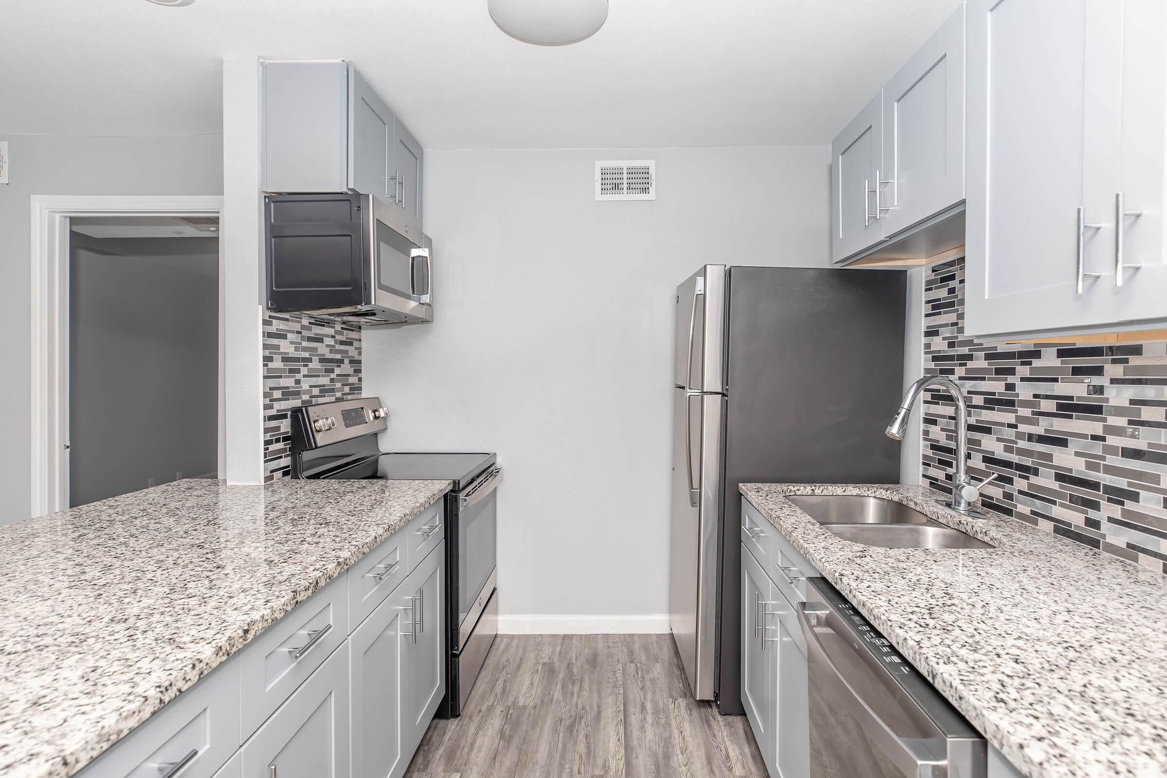 Modern kitchen featuring granite countertops, stainless steel appliances including a stove and refrigerator, gray cabinetry, and a stylish backsplash with black and white tiles. The space is well-lit with overhead lighting and has an open layout.
