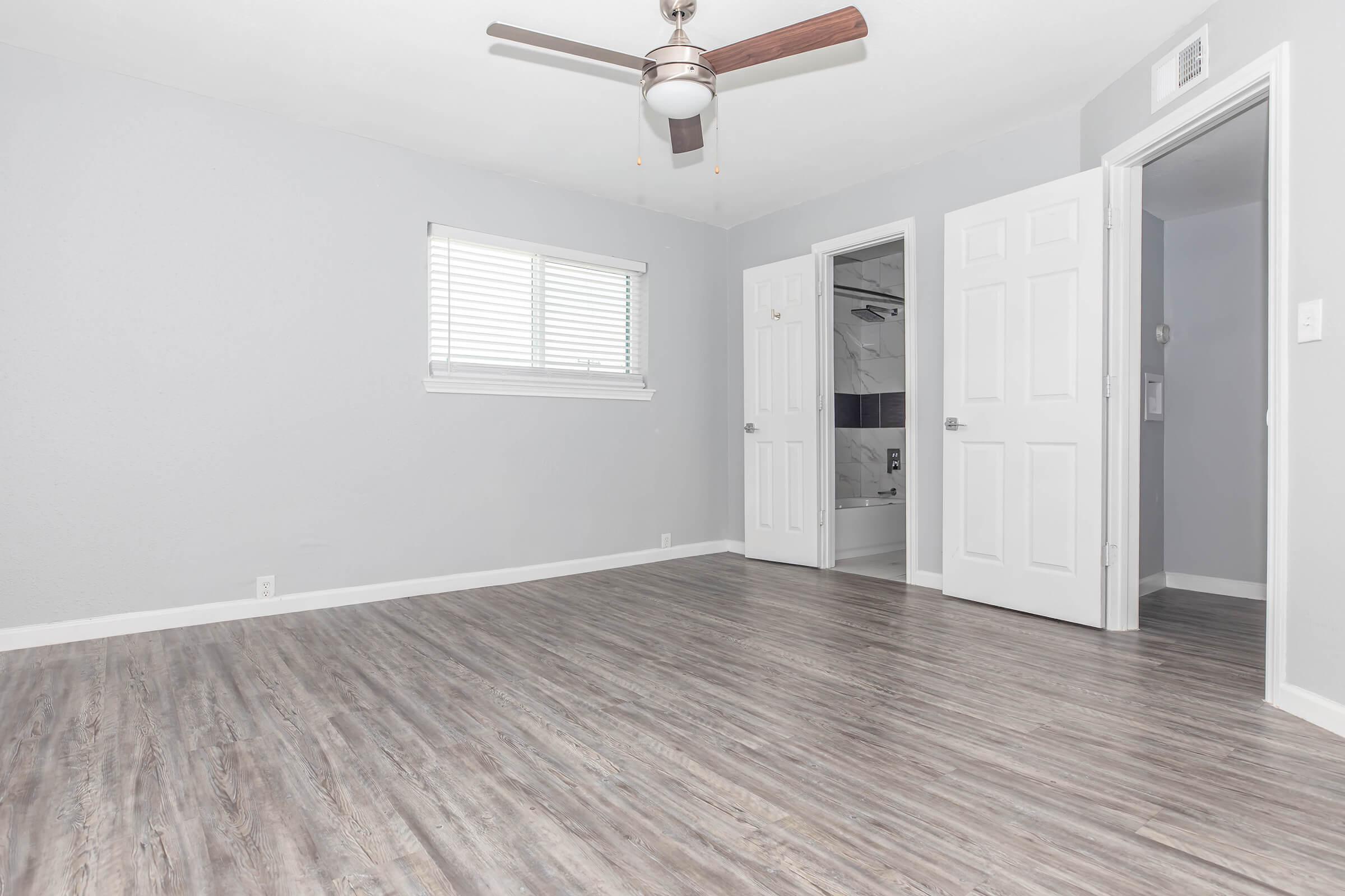 A spacious, empty room featuring light gray walls and a ceiling fan. There are two doors: one leading to a bathroom and the other an entry or closet. The floor is covered with light wood-like laminate. A window with horizontal blinds allows natural light into the space.