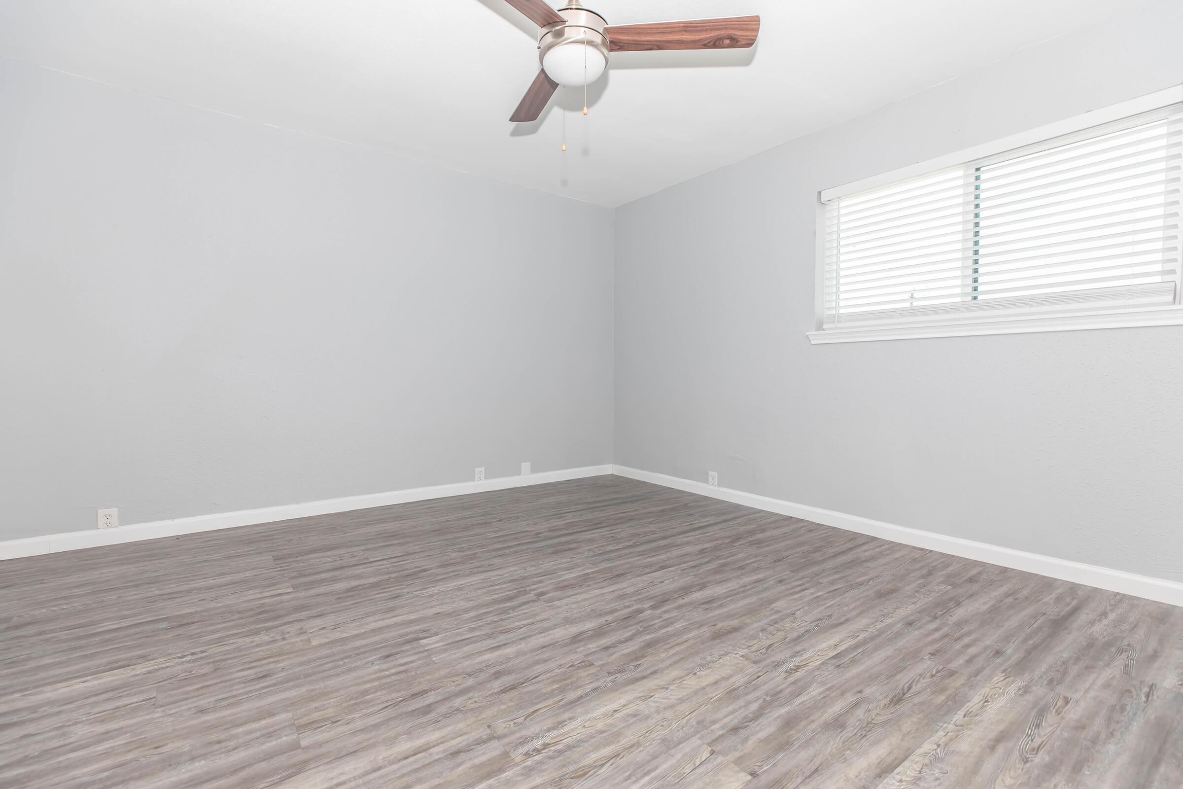 Empty room with gray walls and hardwood-like flooring. A ceiling fan with wooden blades is installed, and there is a window with white blinds letting in natural light. The room features a minimalistic and spacious design, suitable for various purposes.