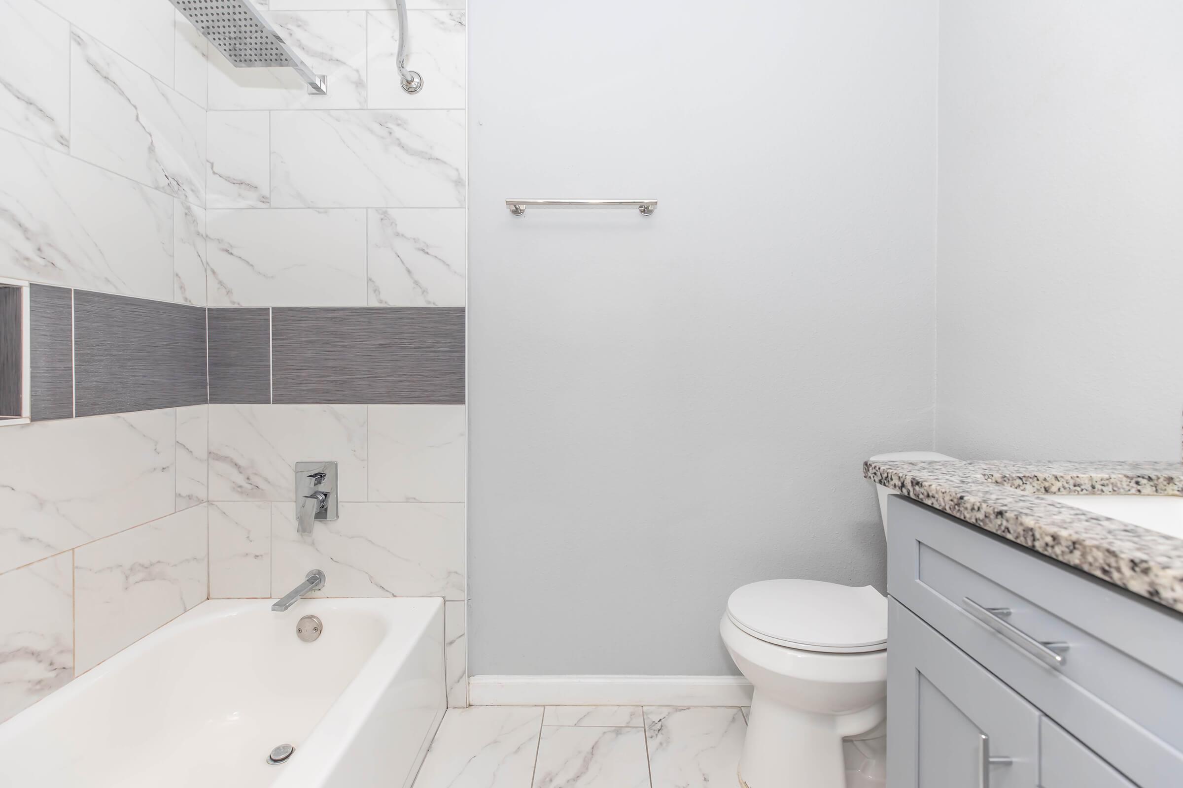 Modern bathroom featuring a white bathtub with a marble surround, a sleek toilet, and light gray cabinetry. The walls are decorated with white and gray marble tiles, creating a clean and contemporary aesthetic. A towel bar is mounted on the wall, and a granite countertop is visible.