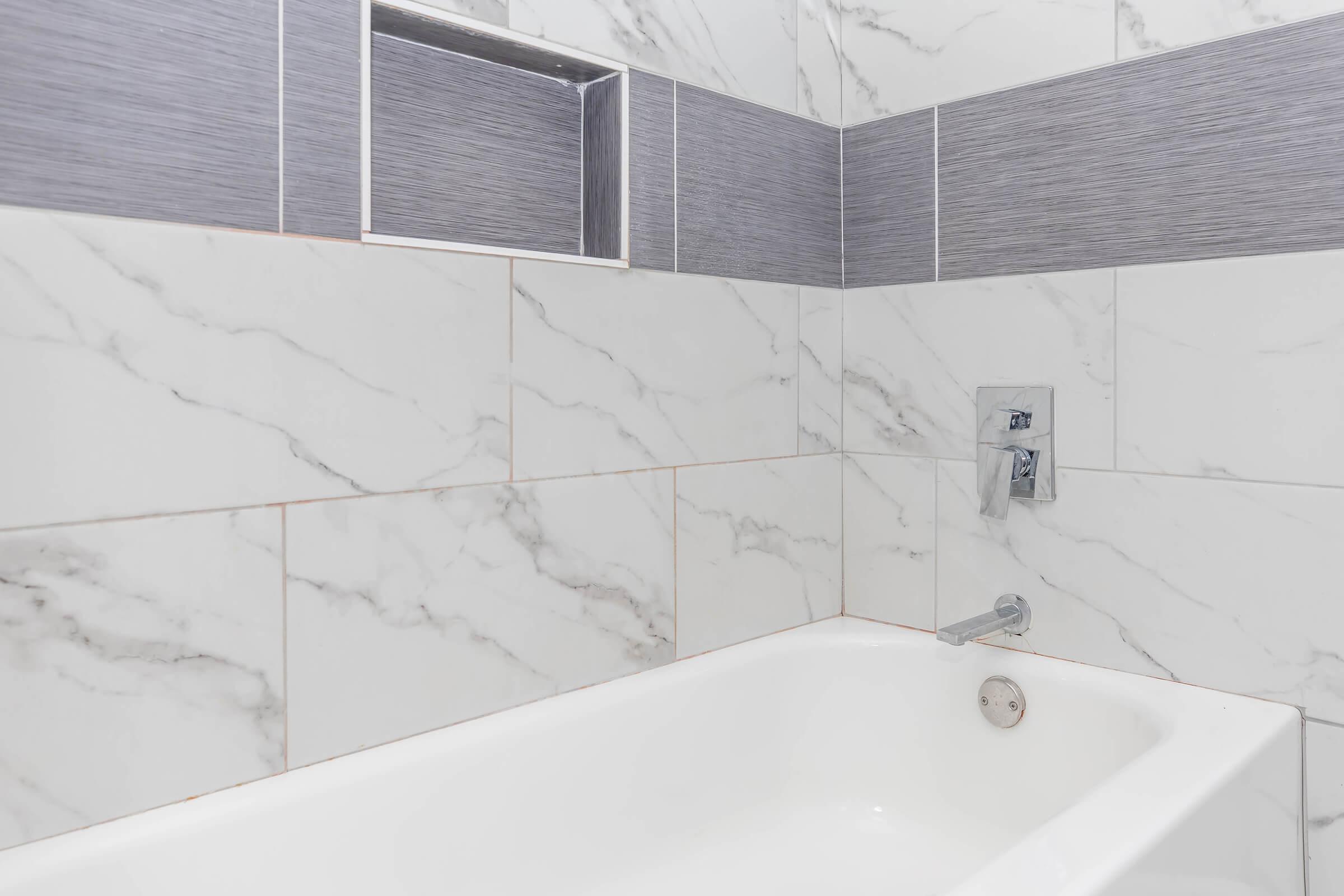 A modern bathroom featuring a white bathtub against a backdrop of elegant marble tiles. The walls are adorned with a mix of white and gray marble patterns, and there is a sleek faucet installed beside the tub. A recessed shelf is built into the wall for storage or decor.