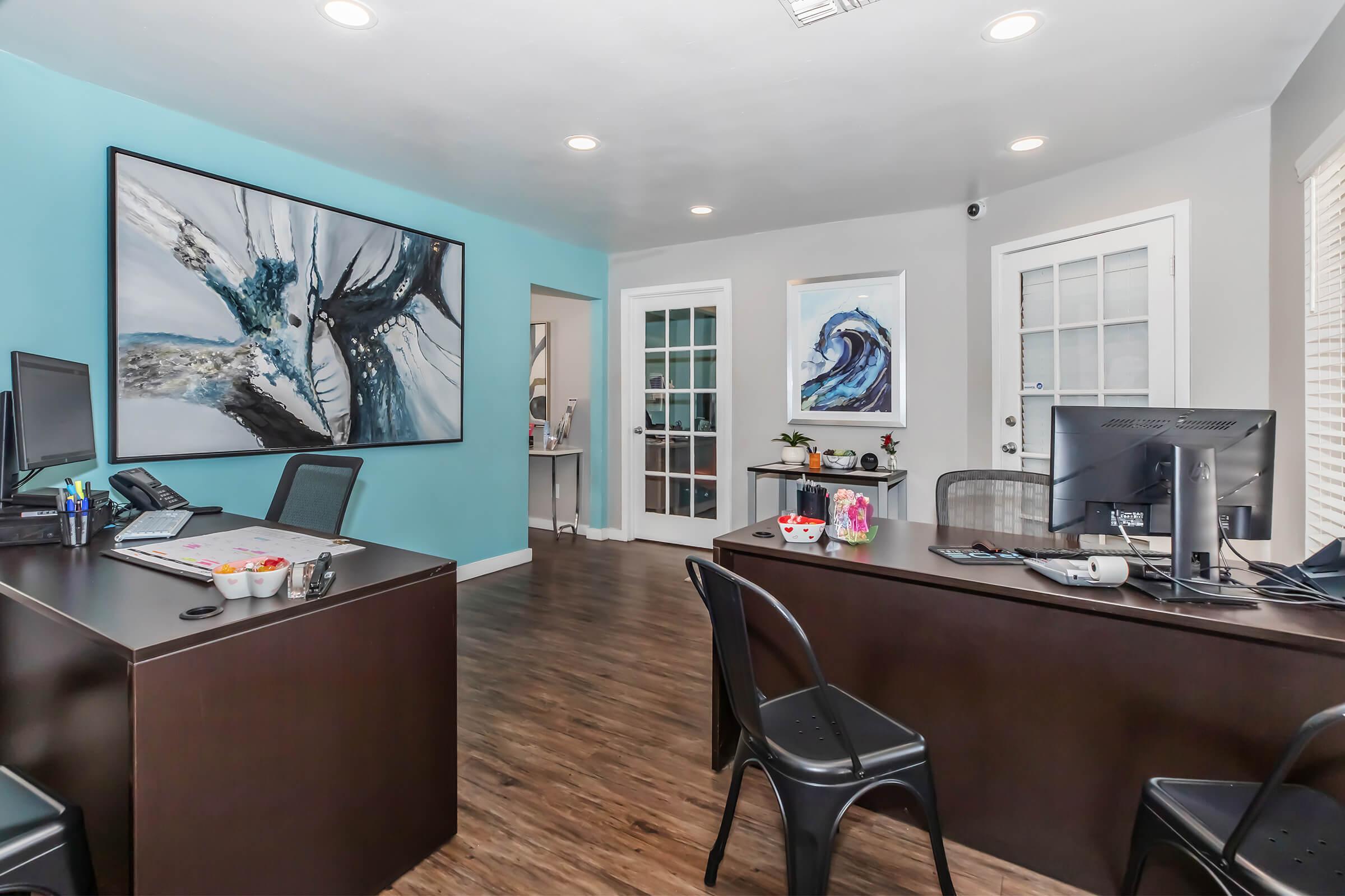 An office interior featuring two dark wood desks with computers, black chairs, and a light blue wall. One wall has a large abstract painting, while another has two smaller framed artworks. There is a door with glass panels in the background, and the floor is made of a wood-like material.