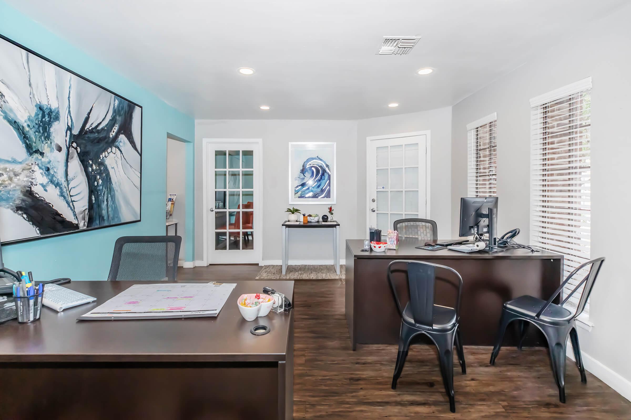 A modern office space featuring two desks with black chairs. One desk has a whiteboard and office supplies. The walls are painted in soft colors, with large artwork on one wall and a wave-themed print on another. Large windows provide natural light, and the flooring is dark wood.
