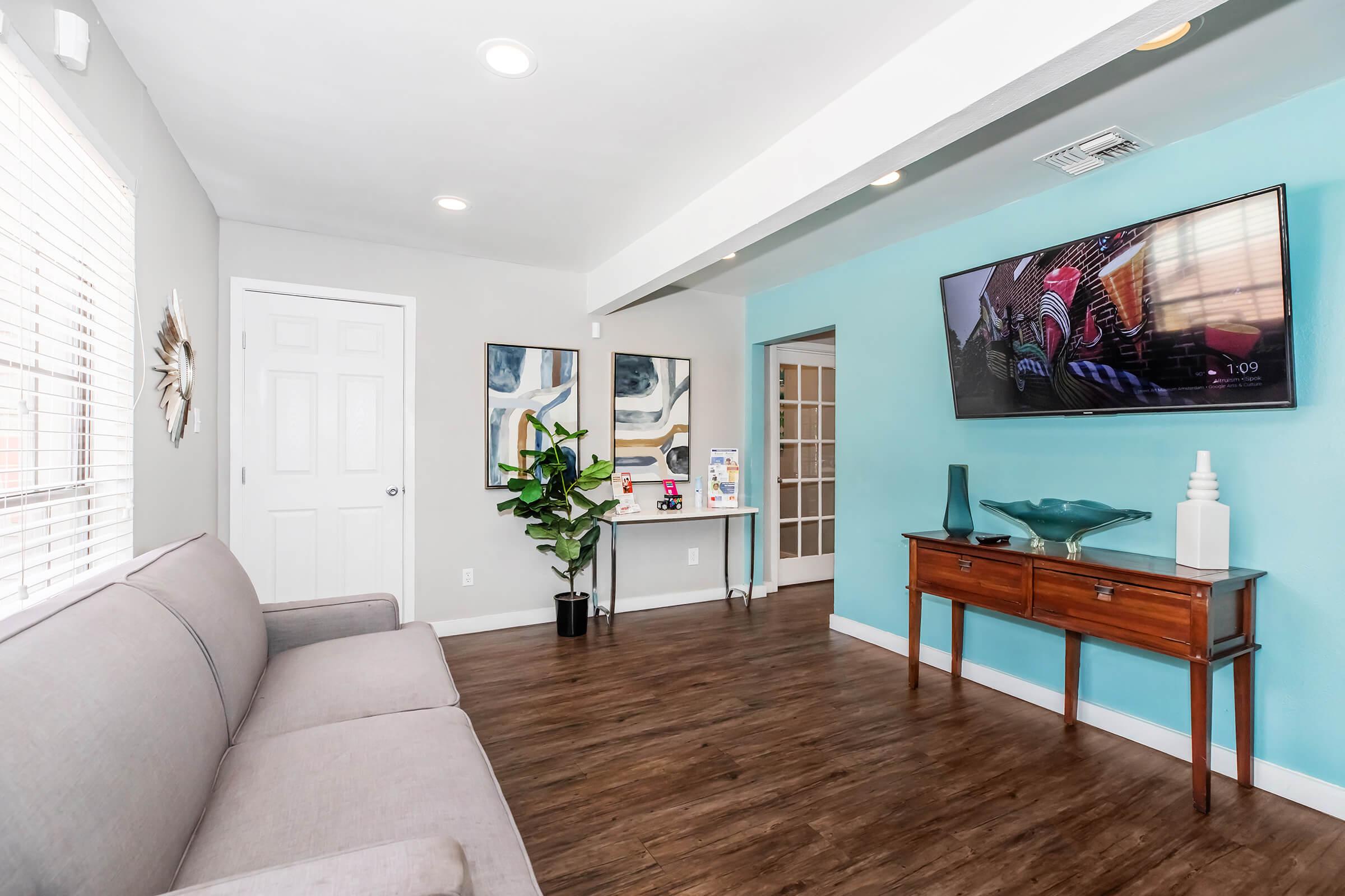 A modern living room featuring a gray sofa, a turquoise accent wall with a mounted television, and wooden flooring. A decorative plant in the corner adds a touch of greenery. A wooden side table displays a blue decorative bowl, and there are framed art pieces on the wall. A door leads to another room.