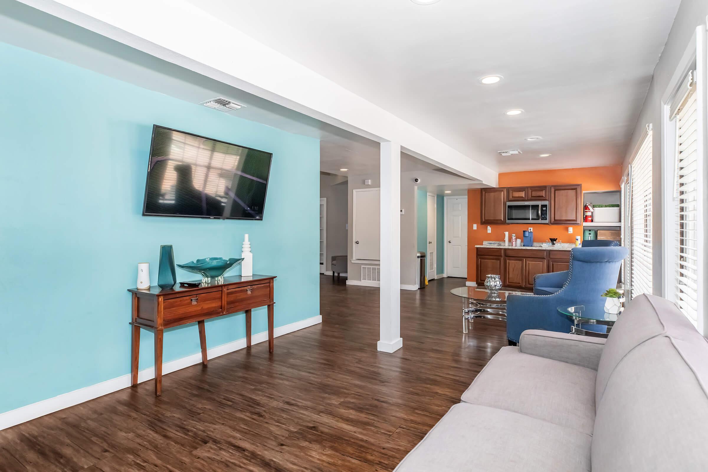 A cozy living room featuring a light gray sofa, a wooden console table with a decorative piece, and a large flat-screen TV mounted on a blue wall. In the background, there's an open kitchen with wooden cabinetry and an orange accent wall. Natural light floods the space through the windows, creating a welcoming ambiance.