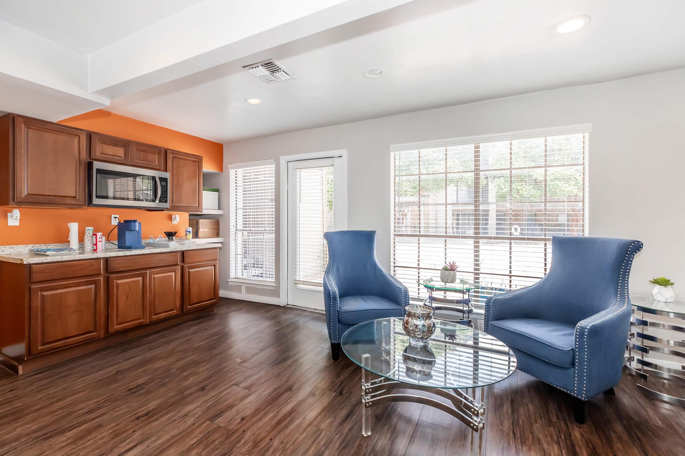 Bright and modern living space featuring a cozy seating area with two blue armchairs flanking a glass coffee table. The kitchen area is visible in the background, showcasing wooden cabinets and a countertop. Large windows provide natural light, with a view of an outdoor patio area.
