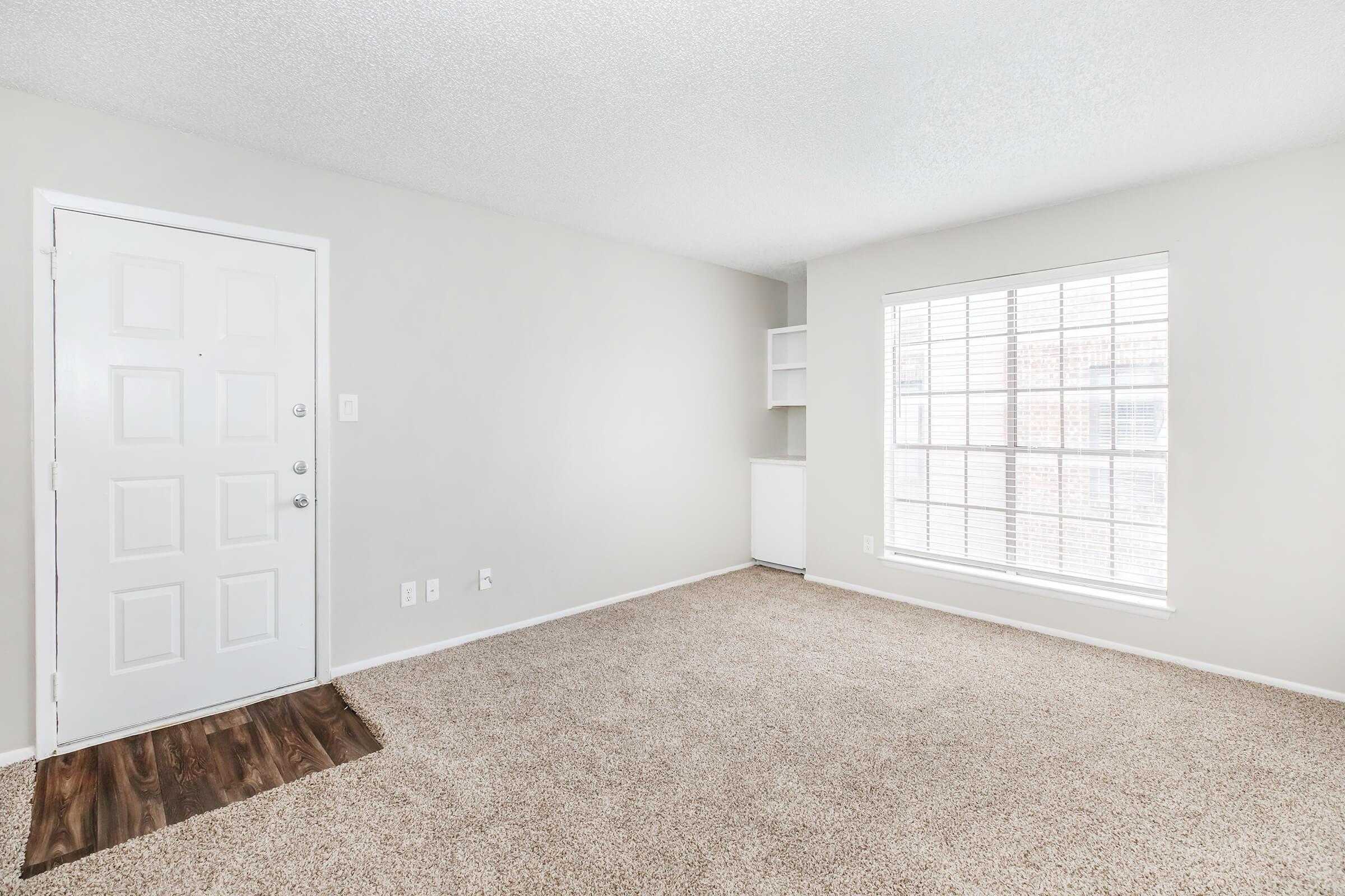A brightly lit living room with light-colored walls and a large window featuring white blinds. The floor is covered in beige carpet, and there is a simple white door on the left. A small shelf is visible beside the window, creating a cozy and inviting atmosphere.