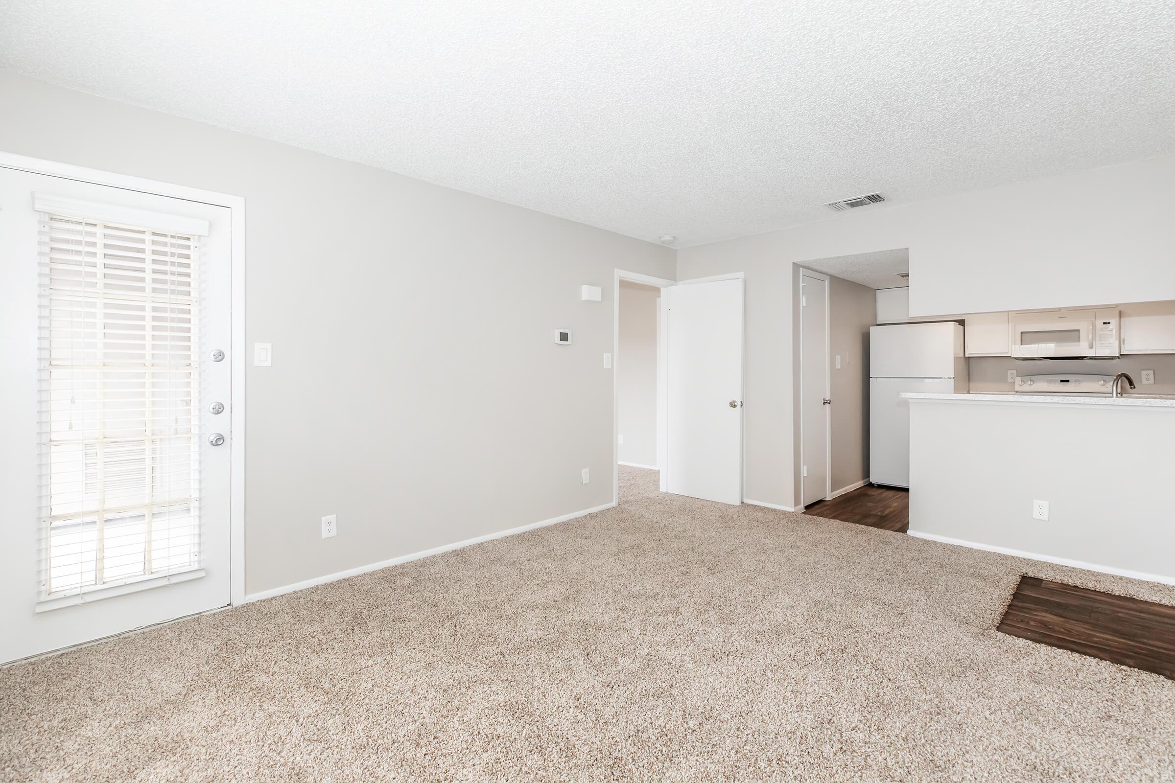 Bright and airy interior of an apartment featuring a spacious carpeted living area, a door with vertical blinds leading outside, and a view of a kitchen area with white appliances. The walls are painted in a light color, enhancing the openness of the space.