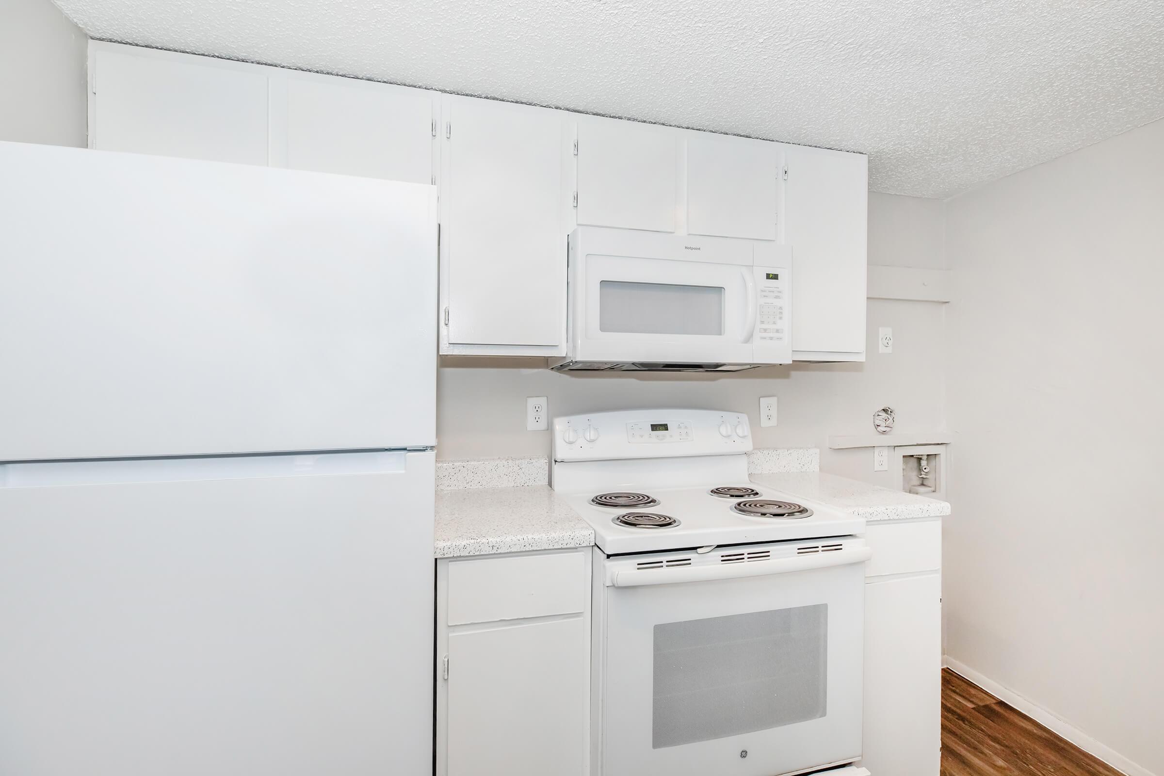 A clean, modern kitchen featuring white cabinetry, a white refrigerator, a white microwave, and an electric stove with four burners. The countertops are light-colored and pristine, and the floor has a wood-like finish, creating a bright and inviting cooking space.