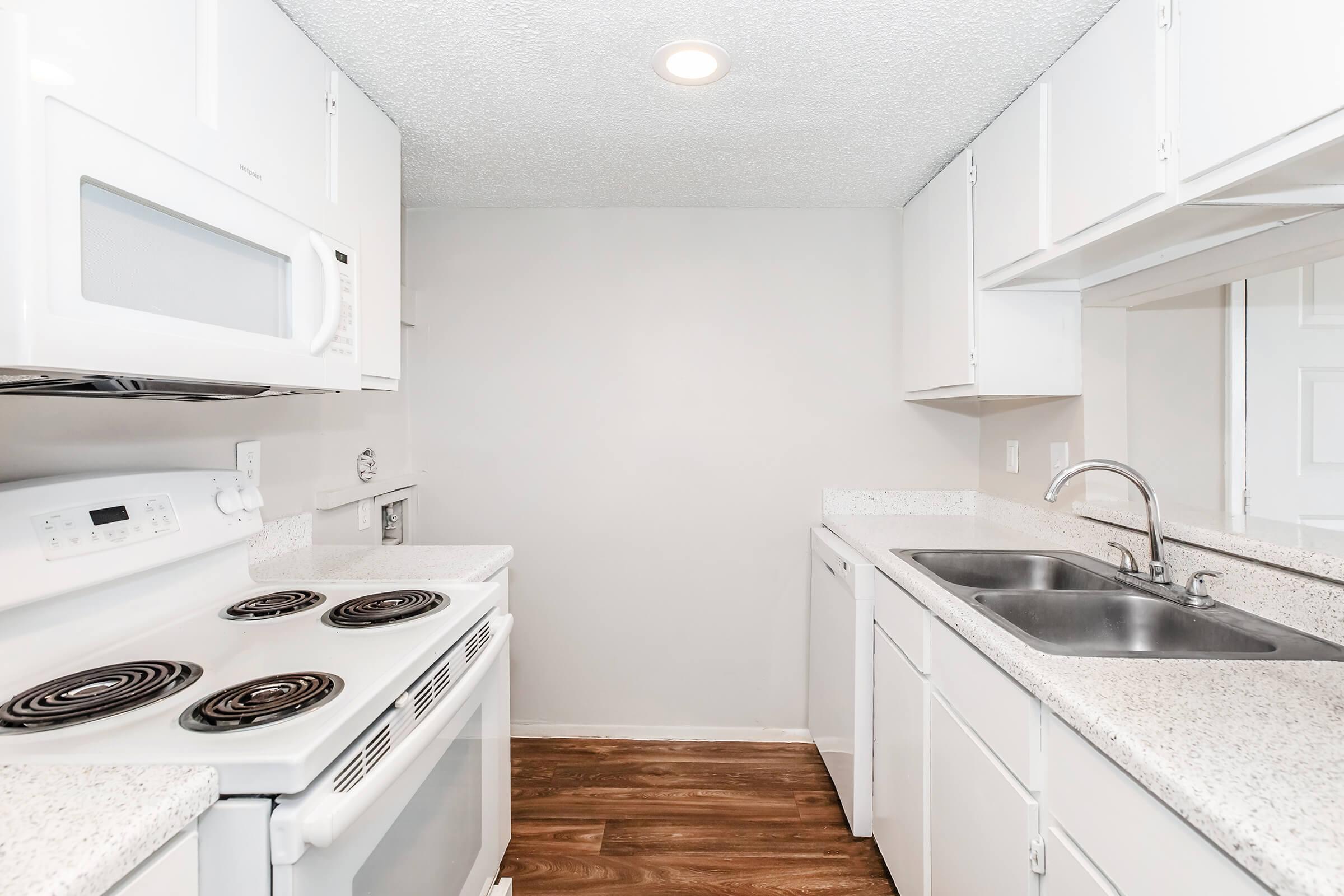 A modern kitchen featuring white cabinets, a double sink, a microwave, and an electric stove with four burners. The countertops are speckled, and the flooring is a light wood laminate. The walls are painted a soft gray, and there is overhead lighting illuminating the space.