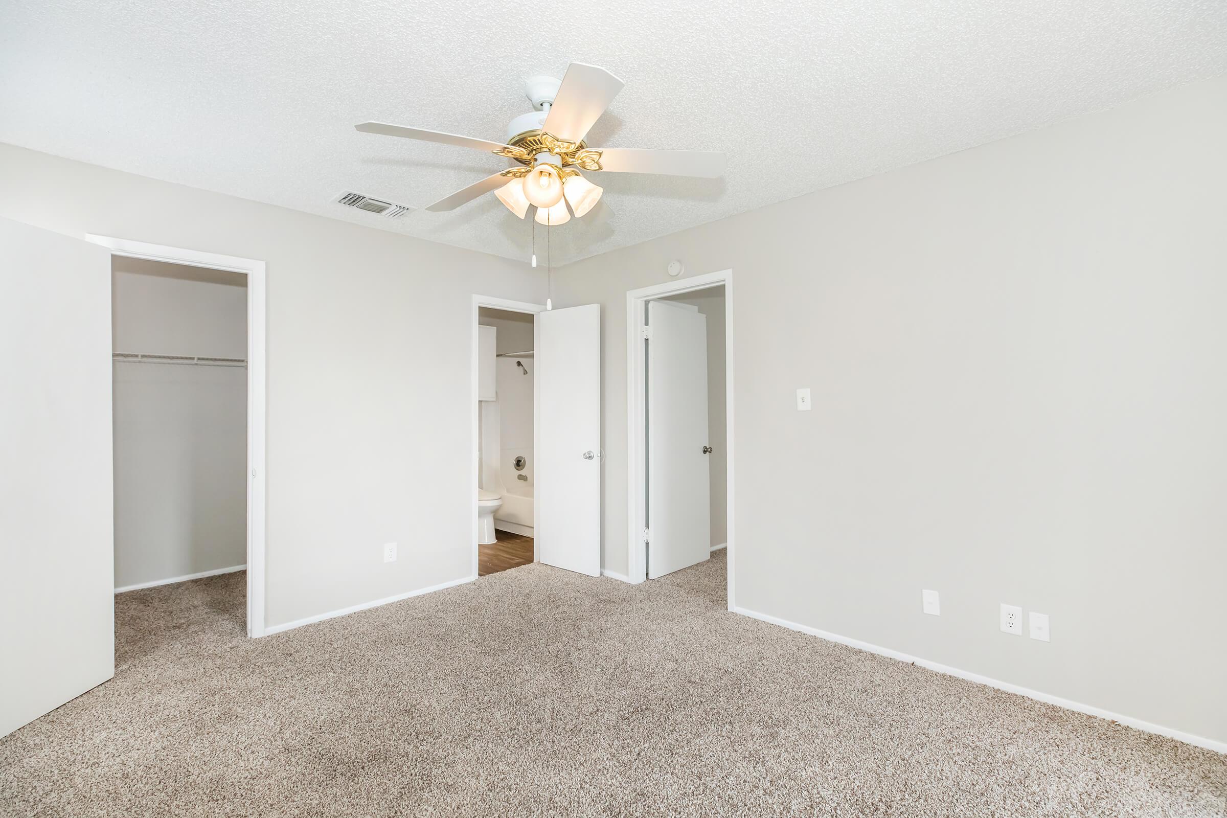 A bright and spacious room featuring light-colored walls and carpet. A ceiling fan is installed, and there are two open doors leading to adjacent areas. One door opens to a closet, while another leads to a bathroom. Natural light enhances the inviting atmosphere of the space.