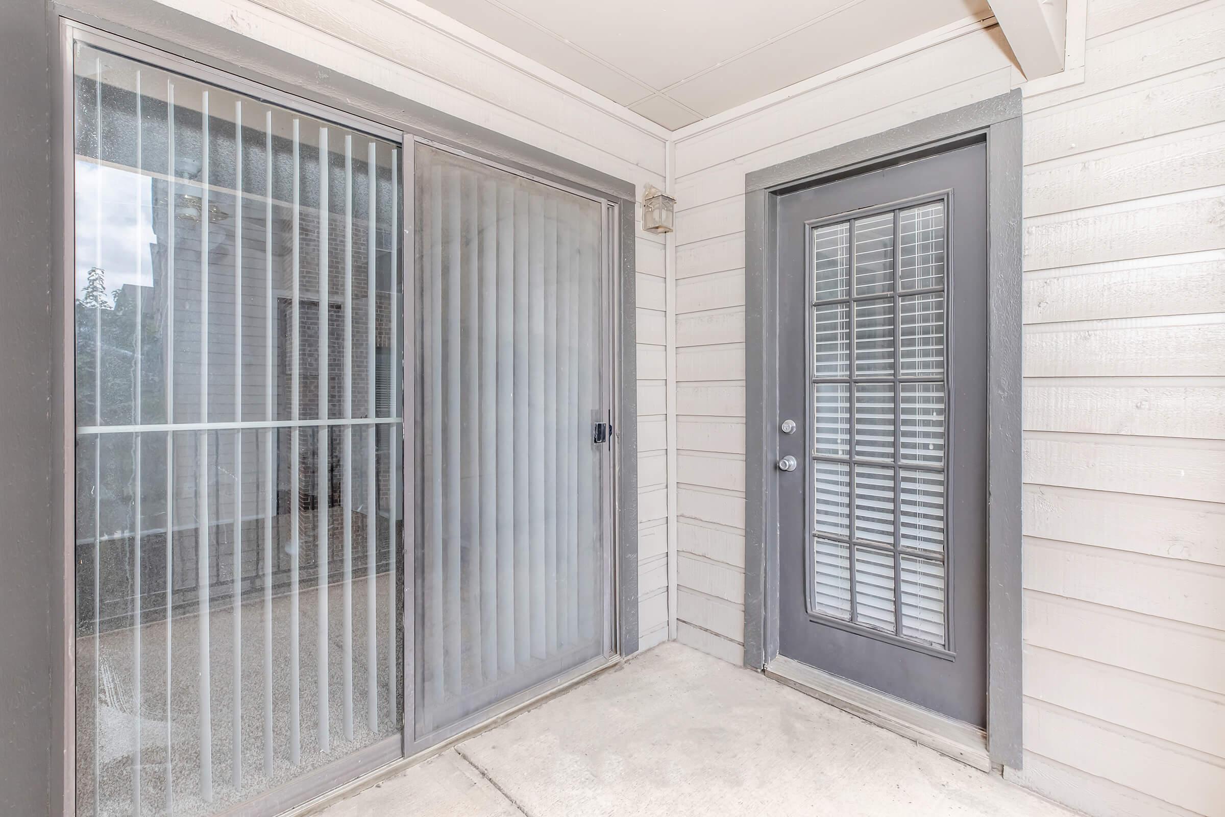 A small entryway featuring a sliding glass door with vertical blinds and a single-pane door with multiple panes of glass. The walls are light-colored wood, and the floor is concrete. Natural light is visible through the glass doors.