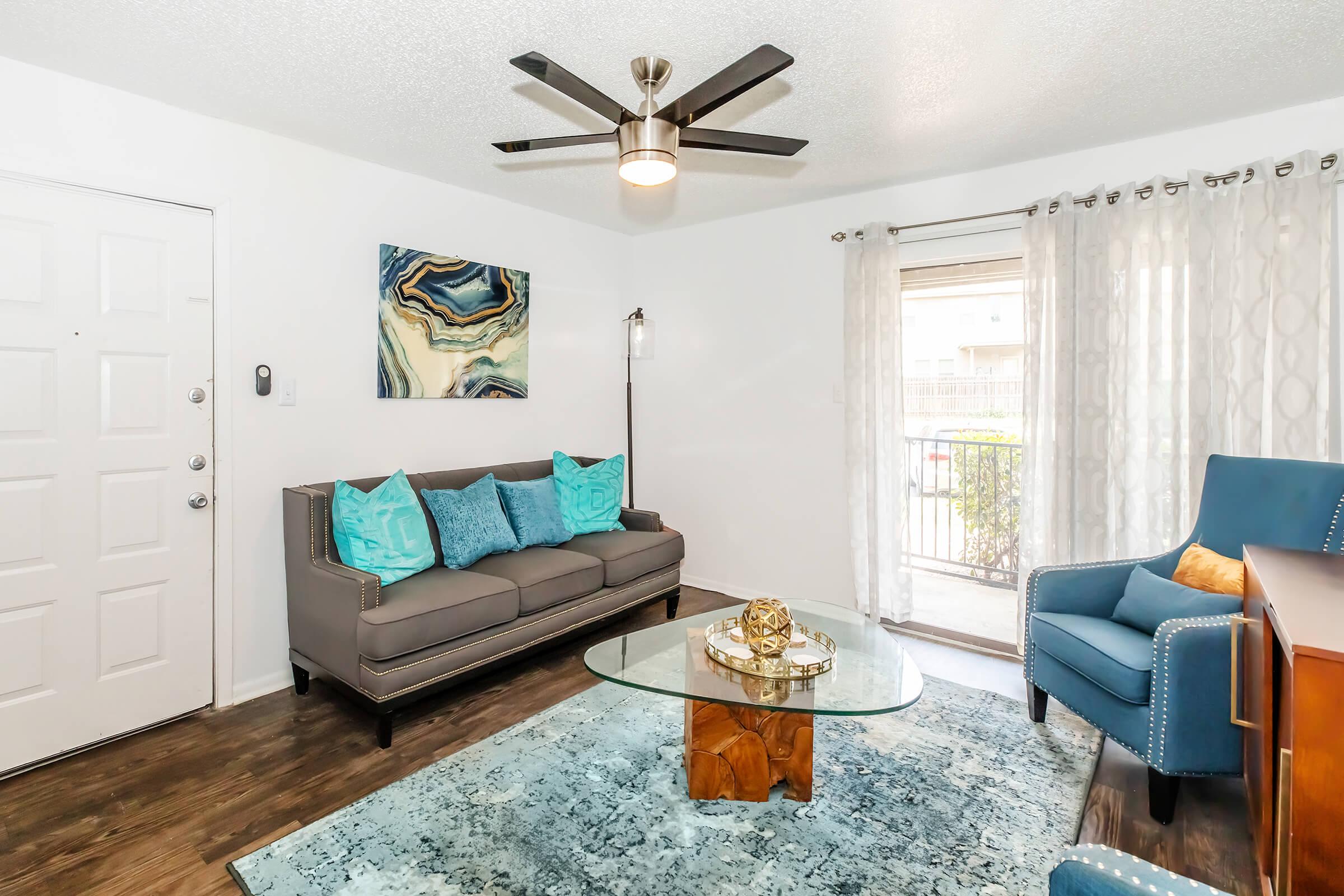 A cozy living room featuring a gray sofa with turquoise accent pillows, a glass coffee table atop a blue area rug, and a blue armchair. Natural light streams in through a window dressed with sheer curtains. A modern ceiling fan and abstract wall art add to the contemporary decor.