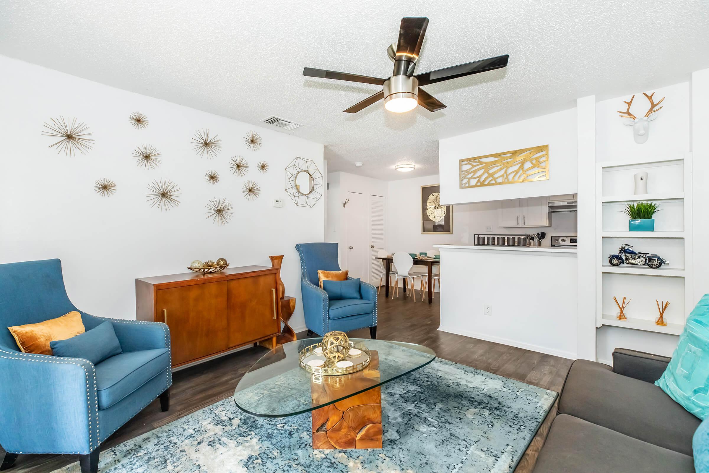 A modern living room featuring two blue armchairs, a glass coffee table on a textured rug, and a wooden sideboard. Decorative wall art and a ceiling fan add character, while a small dining area is visible in the background, showcasing an open concept design.