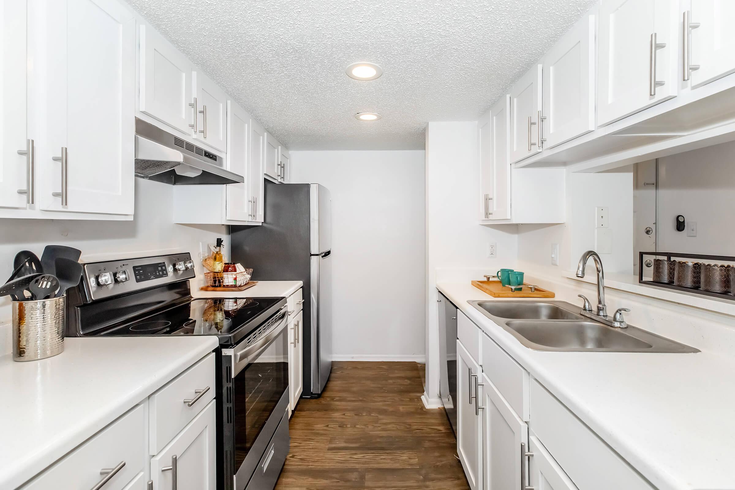 Modern kitchen featuring white cabinetry, stainless steel appliances, and a dual sink. The space includes an oven, stovetop, refrigerator, and ample counter space, with warm wood flooring complementing the bright, clean design.