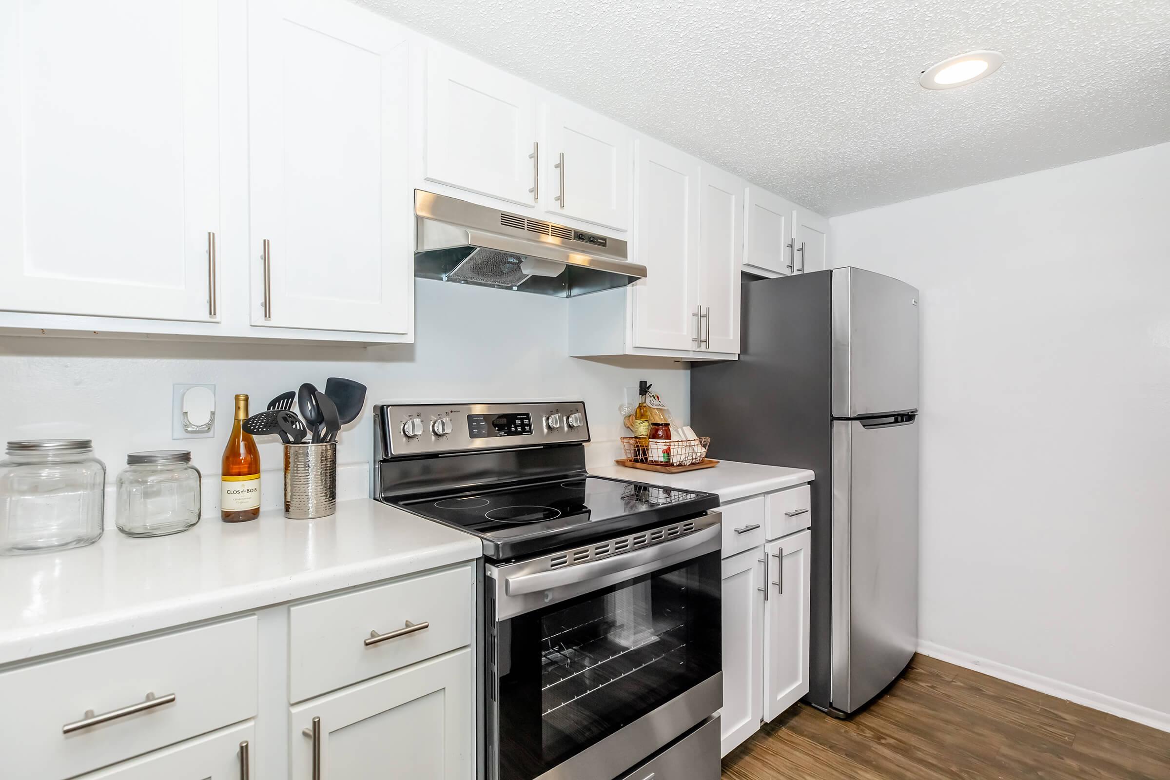 Modern kitchen with white cabinets, a stainless steel refrigerator, and an oven range. Countertop displays glass jars and kitchen utensils, featuring a cozy arrangement with decorative items. The space is well-lit, showcasing clean lines and a contemporary design.