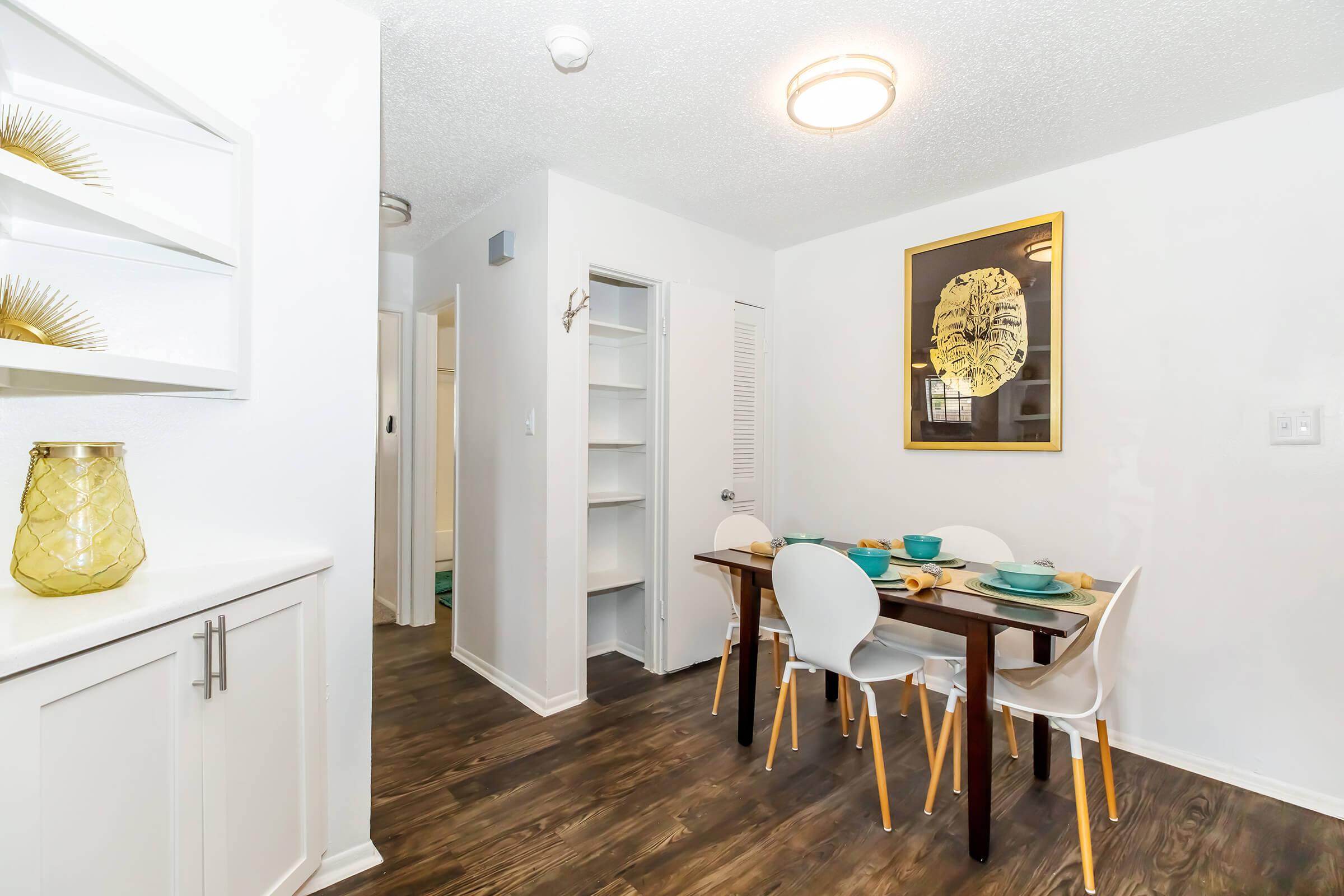 Bright and modern dining area featuring a wooden table set for four with turquoise dishware, surrounded by white chairs. A golden-framed artwork of a lion's face hangs on the wall, and shelves showcase decorative items. The space is warm and inviting, with soft lighting and an open layout.