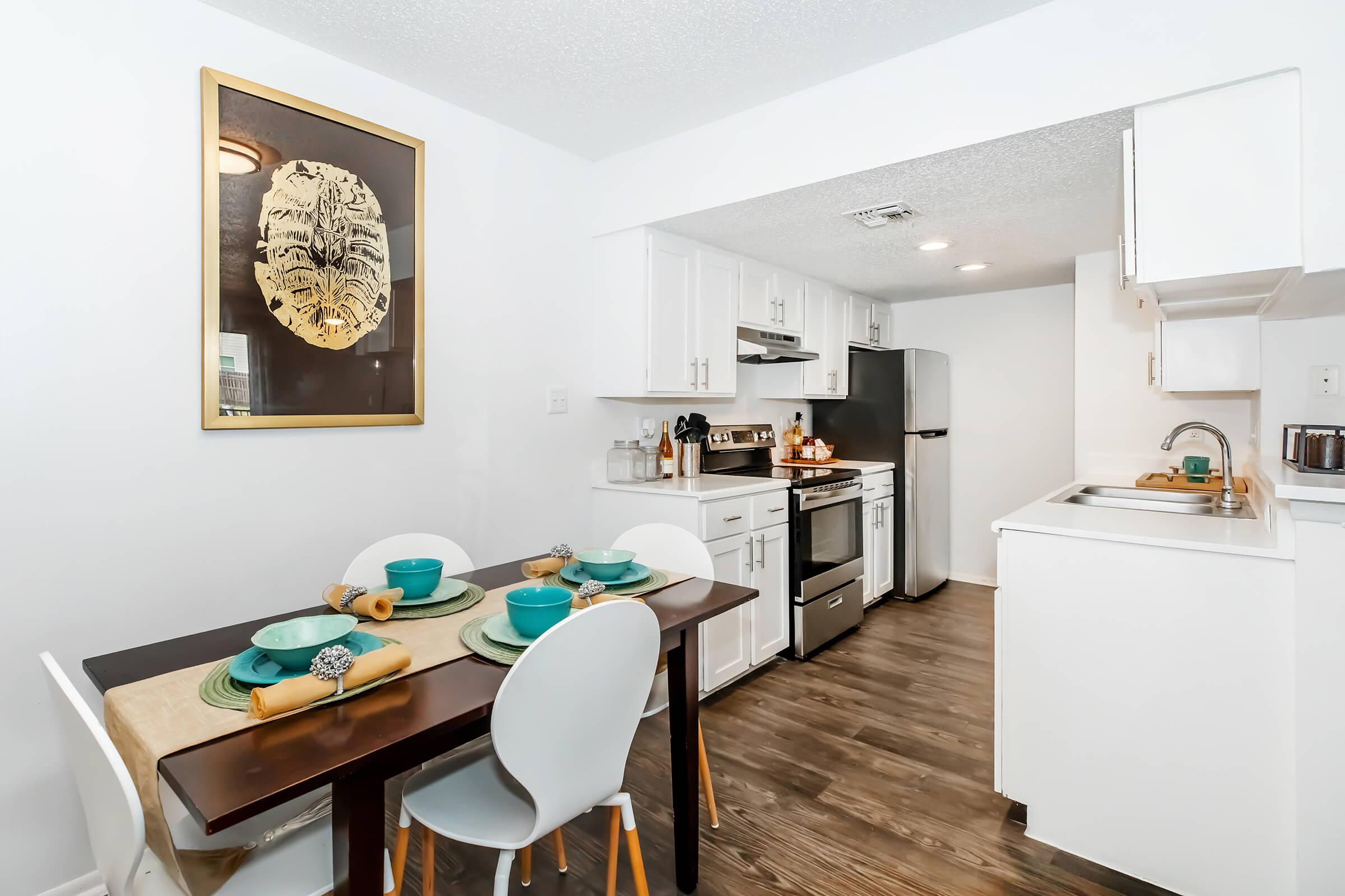A modern kitchen and dining area featuring a white color scheme. The dining table is set with teal plates and complemented by light-colored chairs. A black and gold wall art piece hangs above the table. The kitchen includes stainless steel appliances and white cabinetry, showcasing a clean and inviting space.