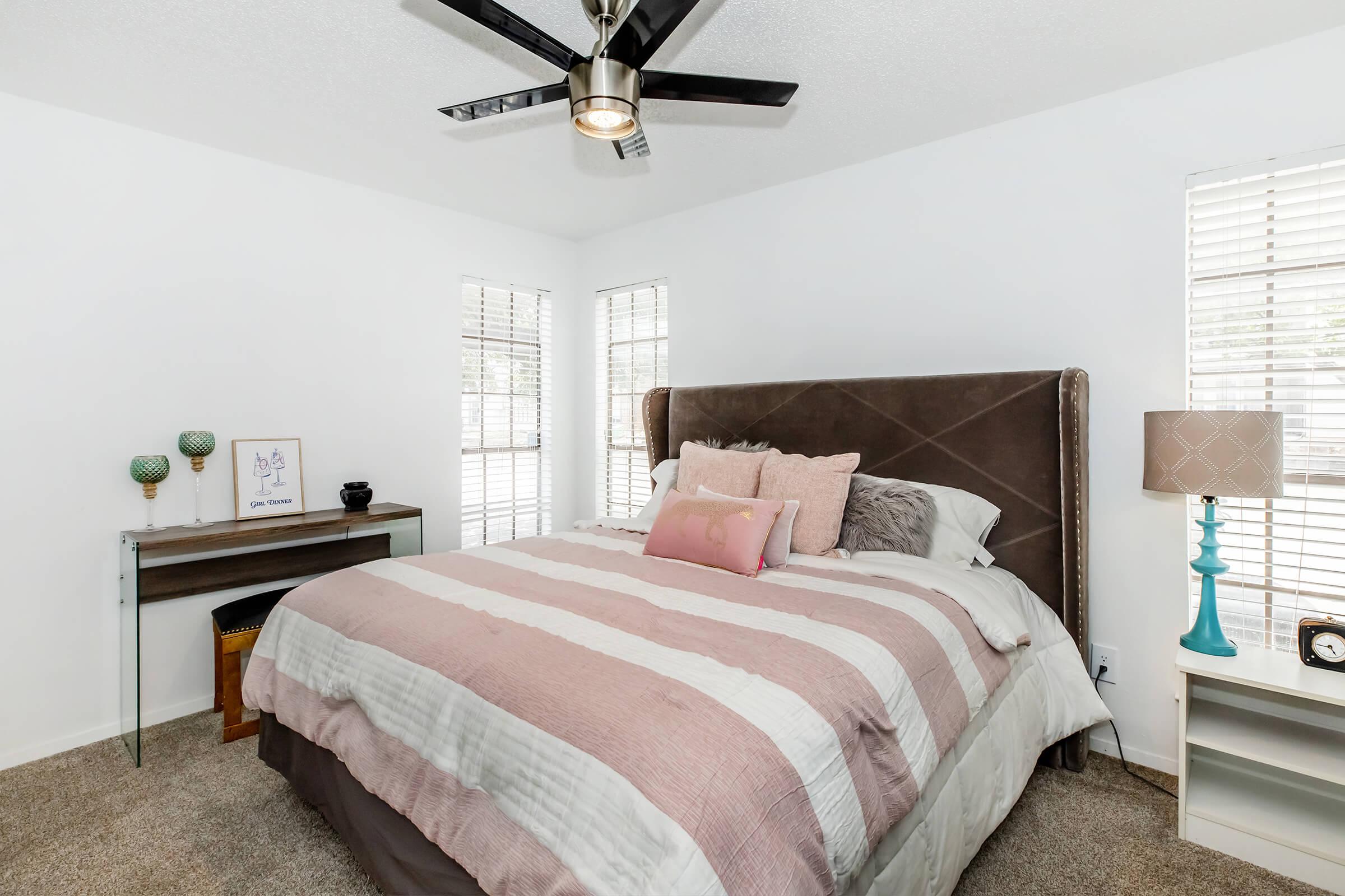 A cozy bedroom featuring a king-sized bed with a striped comforter in pastel colors, accented with decorative pillows. There's a modern ceiling fan, stylish lamps on nightstands, and large windows with blinds. A small desk with a framed picture adds a personal touch to the space.